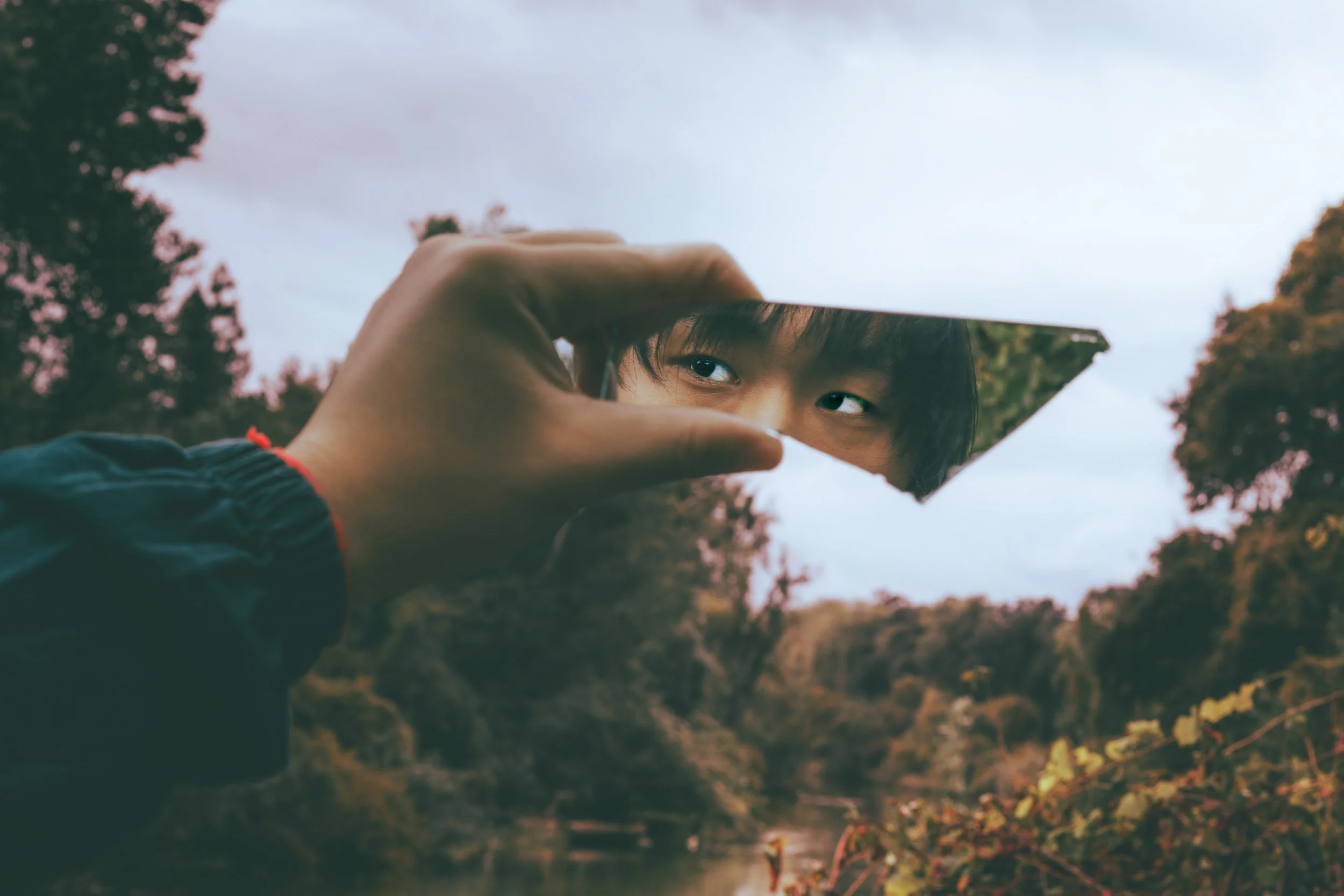 Person holding a broken mirror reflecting their face with their eyes visible, outdoors with trees and a cloudy sky in the background.