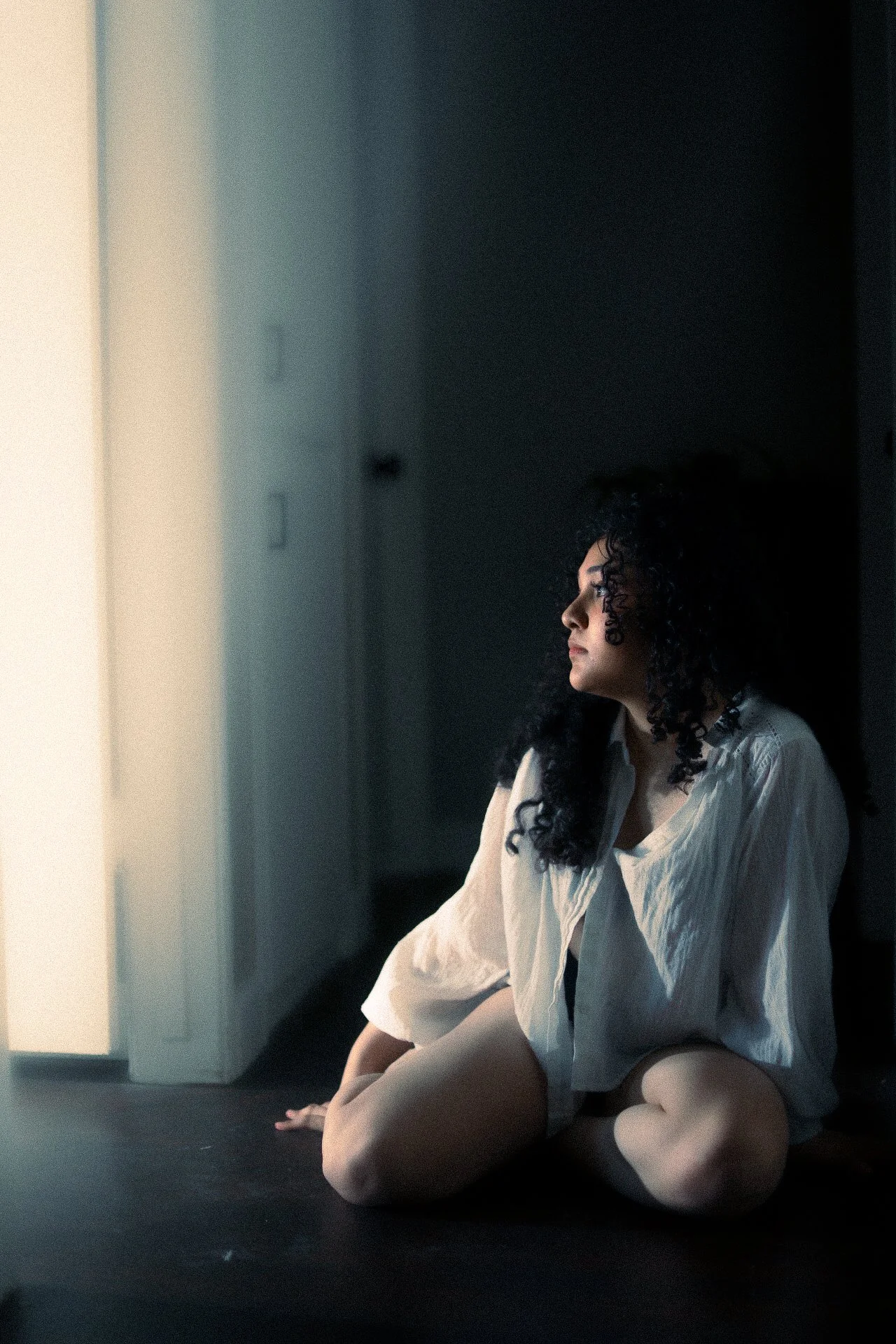 A woman with curly black hair sitting on the floor in a dimly lit room, looking pensively towards a bright light source outside the frame.
