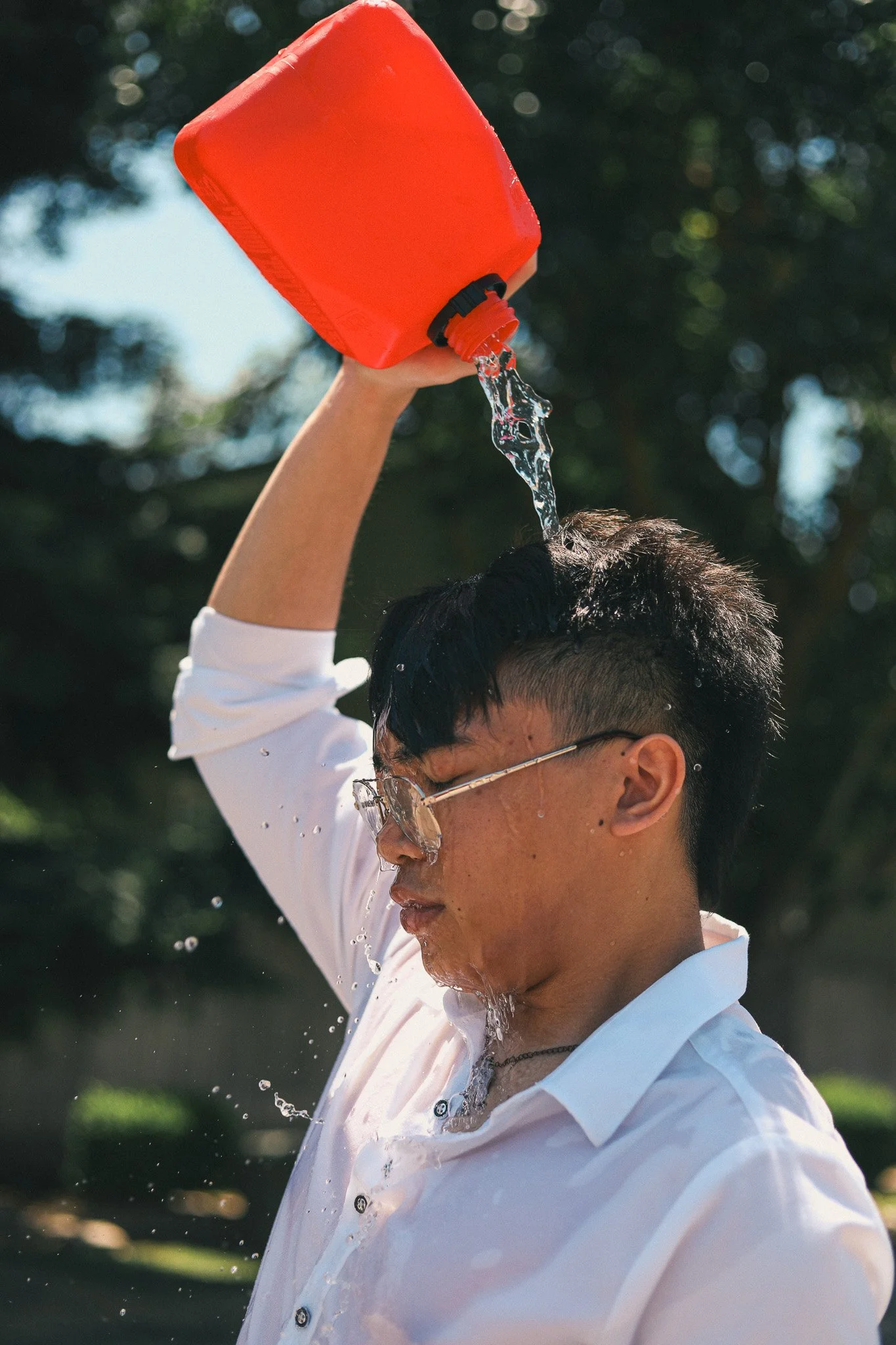 A young man wearing glasses and a white shirt is pouring water over his head from a red container outdoors during daytime.