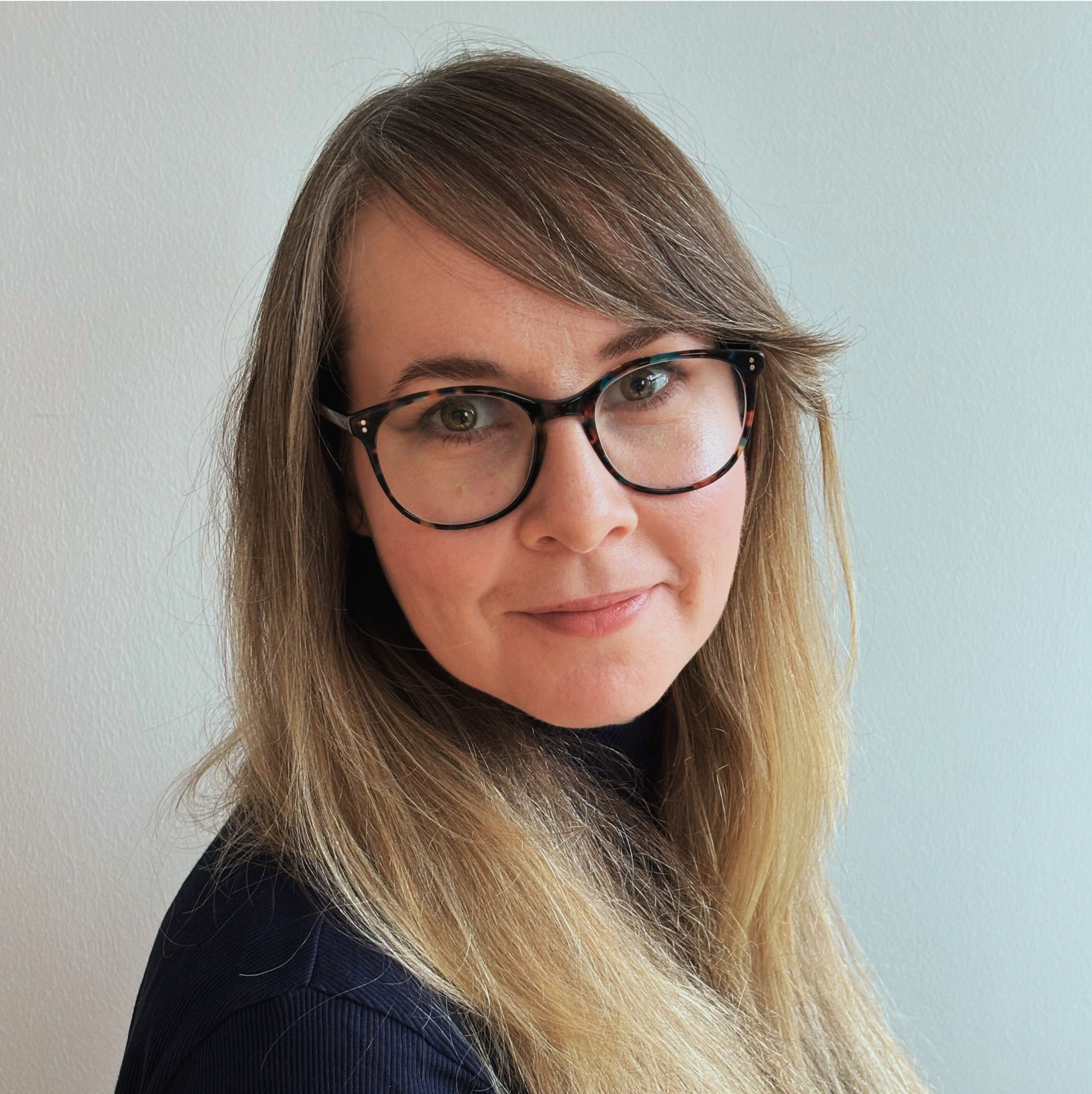 A woman with brown hair wearing black glasses and a black top, smiling slightly, against a plain light-colored background.