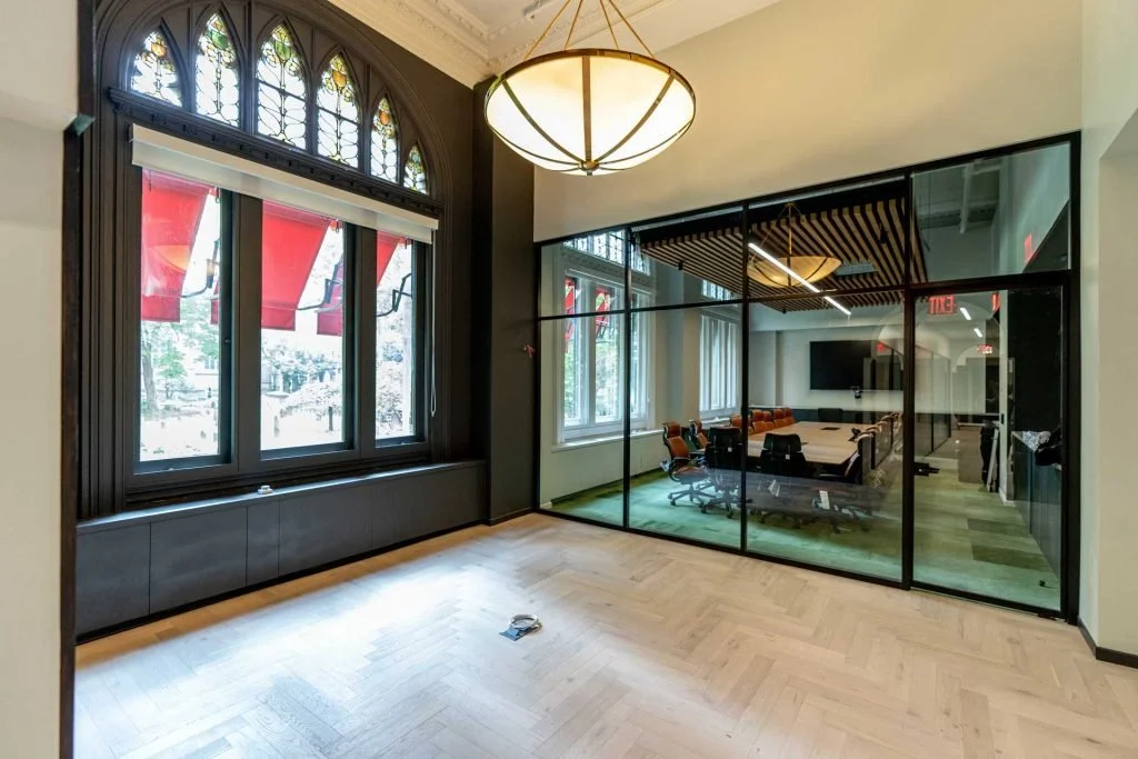 A spacious interior with large stained glass windows and a modern glass-enclosed conference room with a wooden ceiling design, a large meeting table, and chairs, with a chandelier overhead.