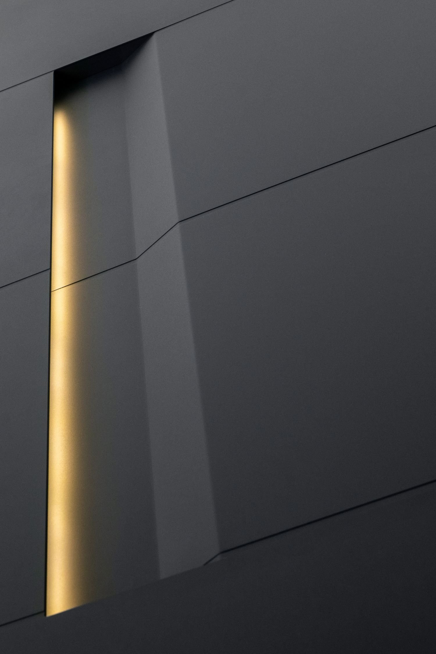 Close-up of a modern, dark gray building with an opening that reveals warm yellow lighting.