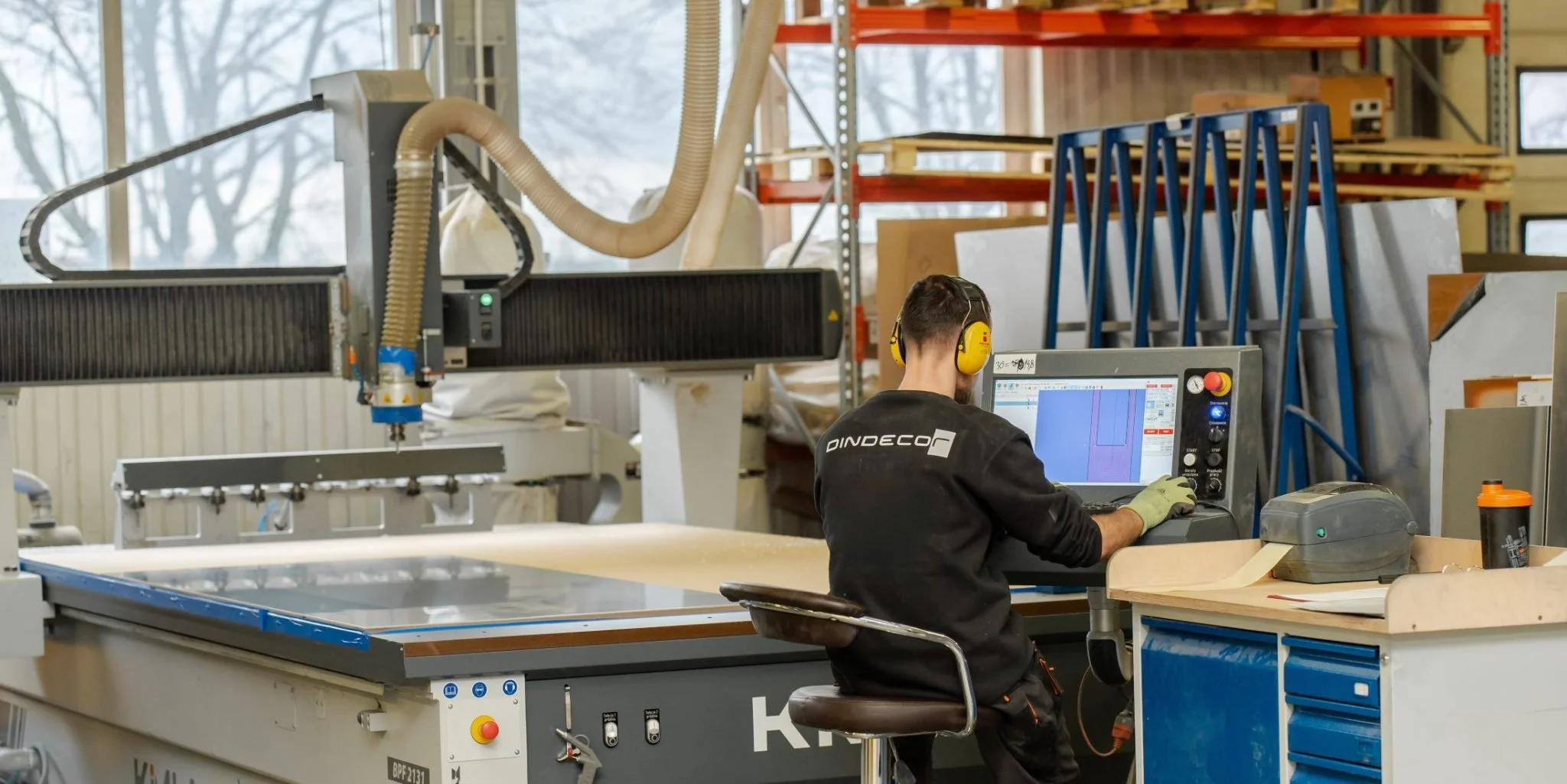 A man wearing yellow ear protection and gloves is working on a computer in a woodworking shop. The shop contains machinery, tools, and shelves with various materials.