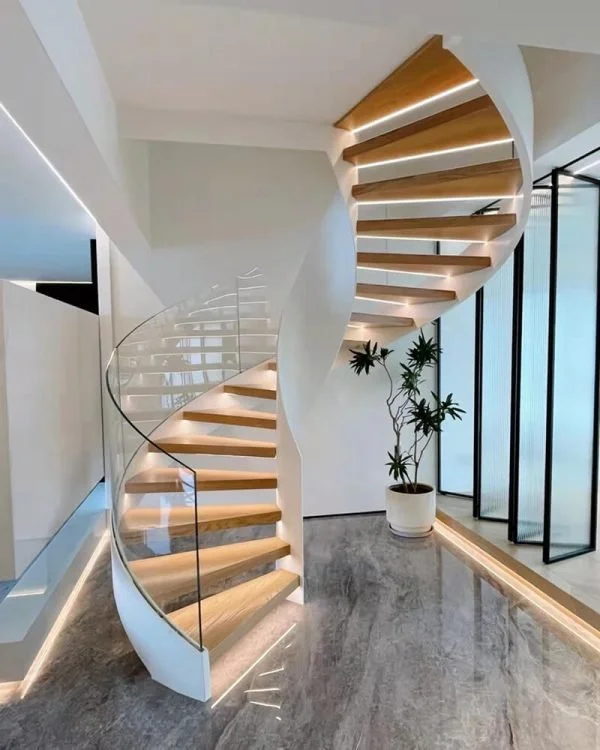 Modern white spiral staircase with wooden steps and glass railing in a bright, minimalist interior with large windows, potted plant, and marble flooring.