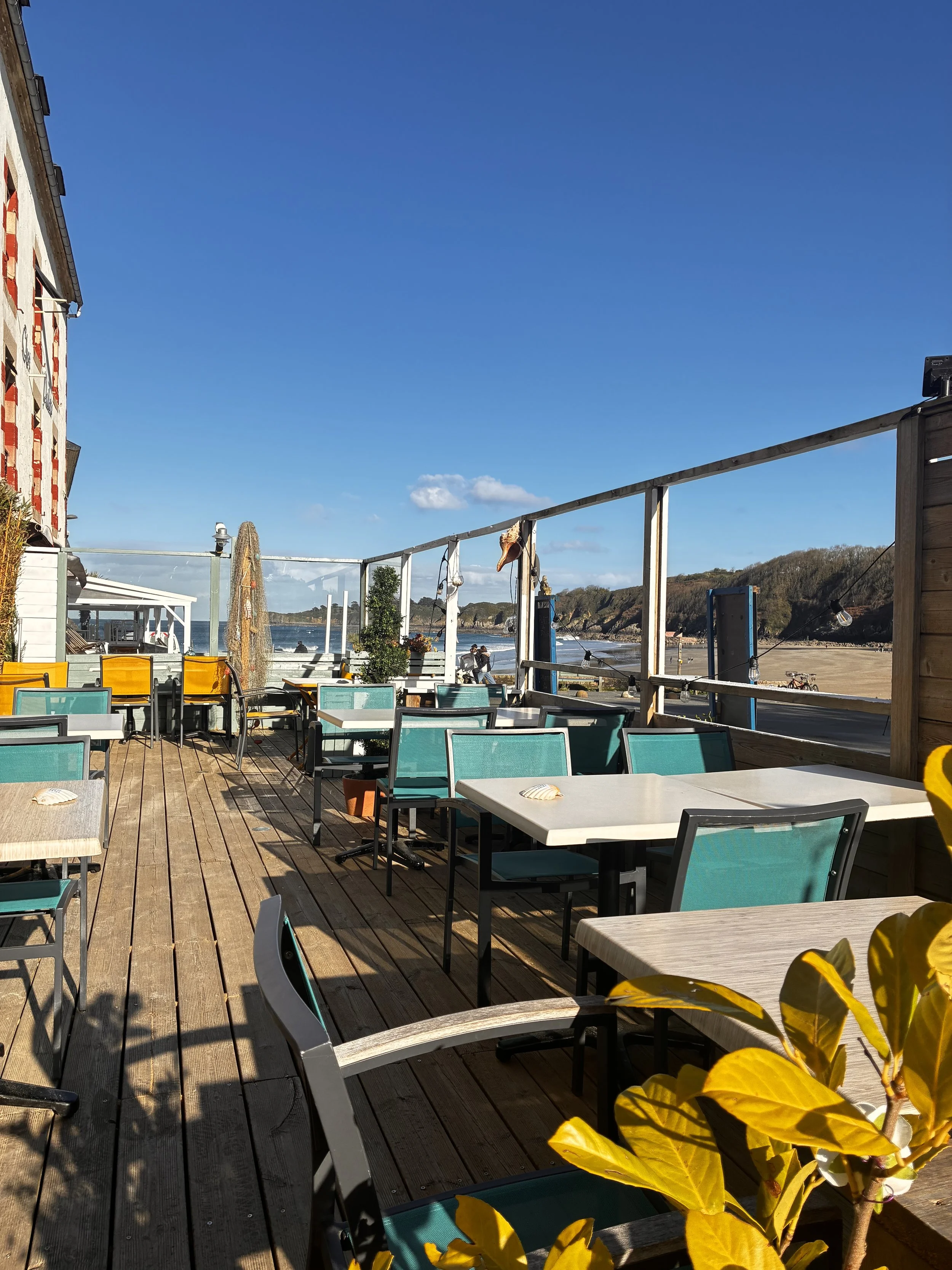 Terrasse extérieure en bois avec des chaises bleues et jaunes, vue sur la plage et la mer, ciel bleu clair, un parasol, des plantes, ambiance ensoleillée