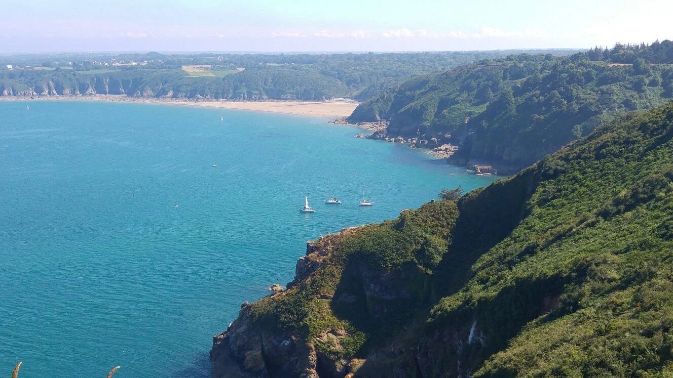 Vue d'une côte avec des falaises vertes et une mer turquoise, avec quelques bateaux à voile et un rivage en arrière-plan.