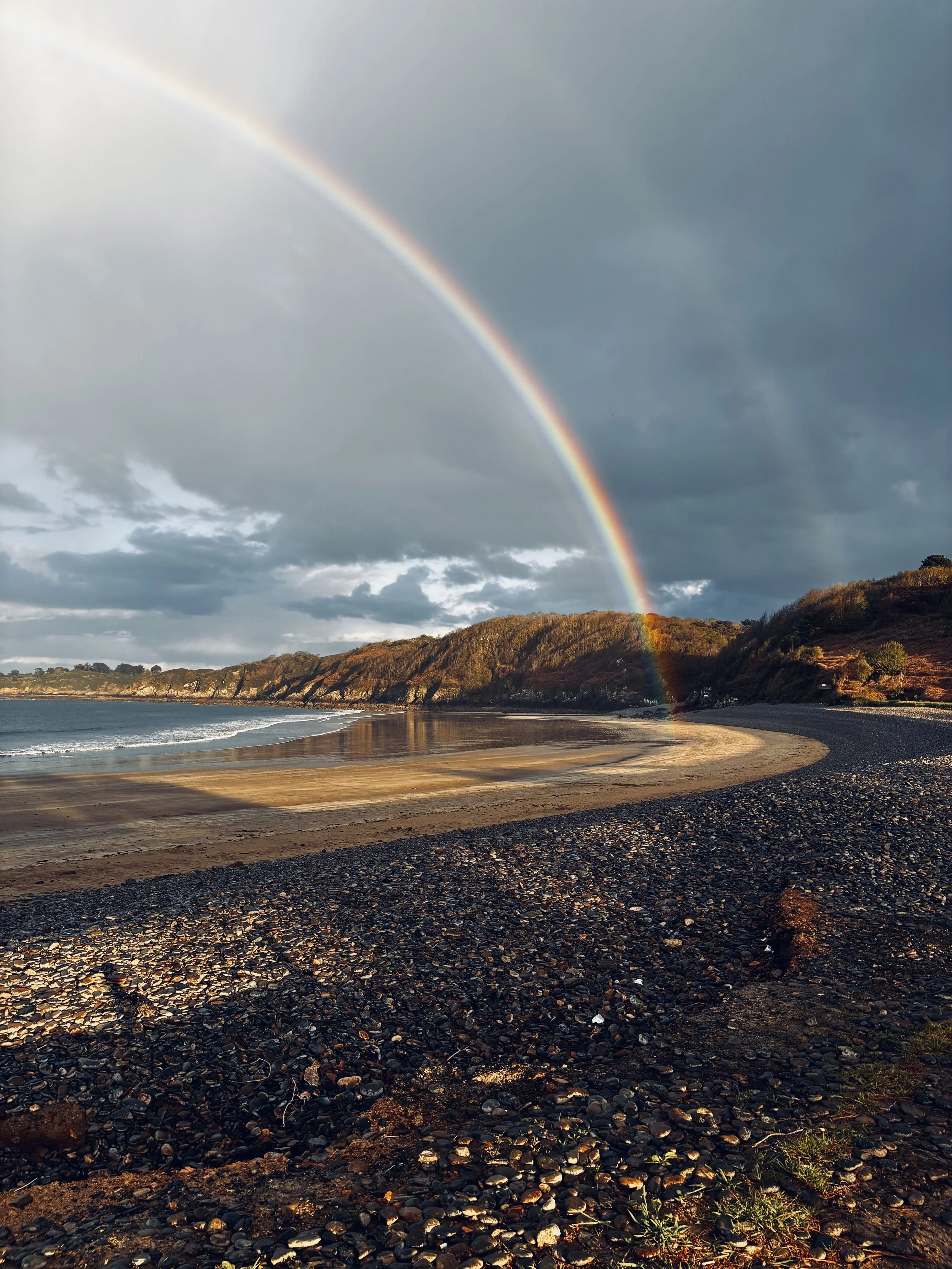 Arc-en-ciel au-dessus d'une plage avec des rochers et des collines en arrière-plan, ciel nuageux.