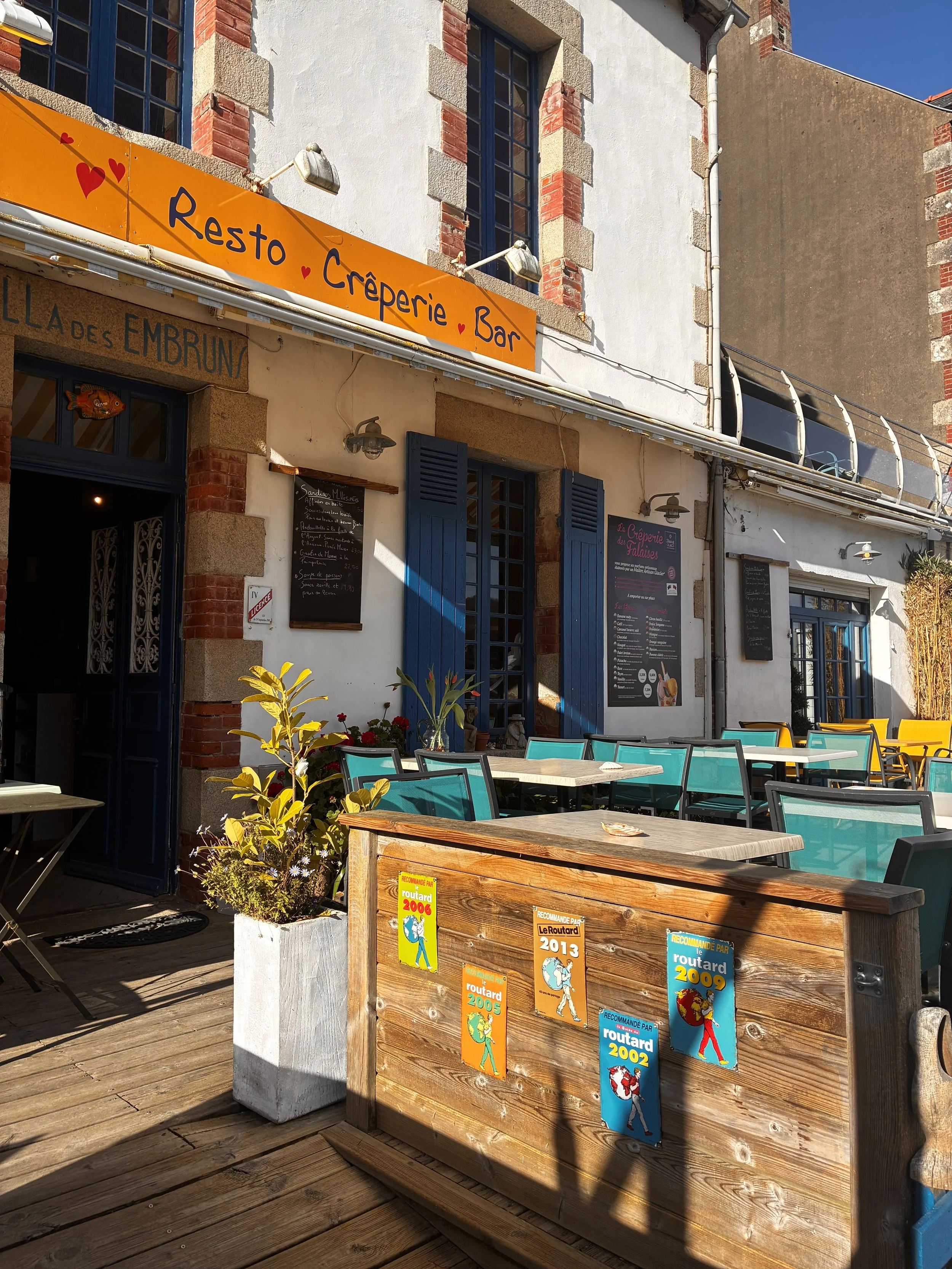 Terrasse extérieure d'un restaurant avec des tables et des chaises colorées, un panneau orange indiquant 'Resto Crêperie Bar', une plante en pot, et des affiches colorées avec des années recommandées par le Routard.