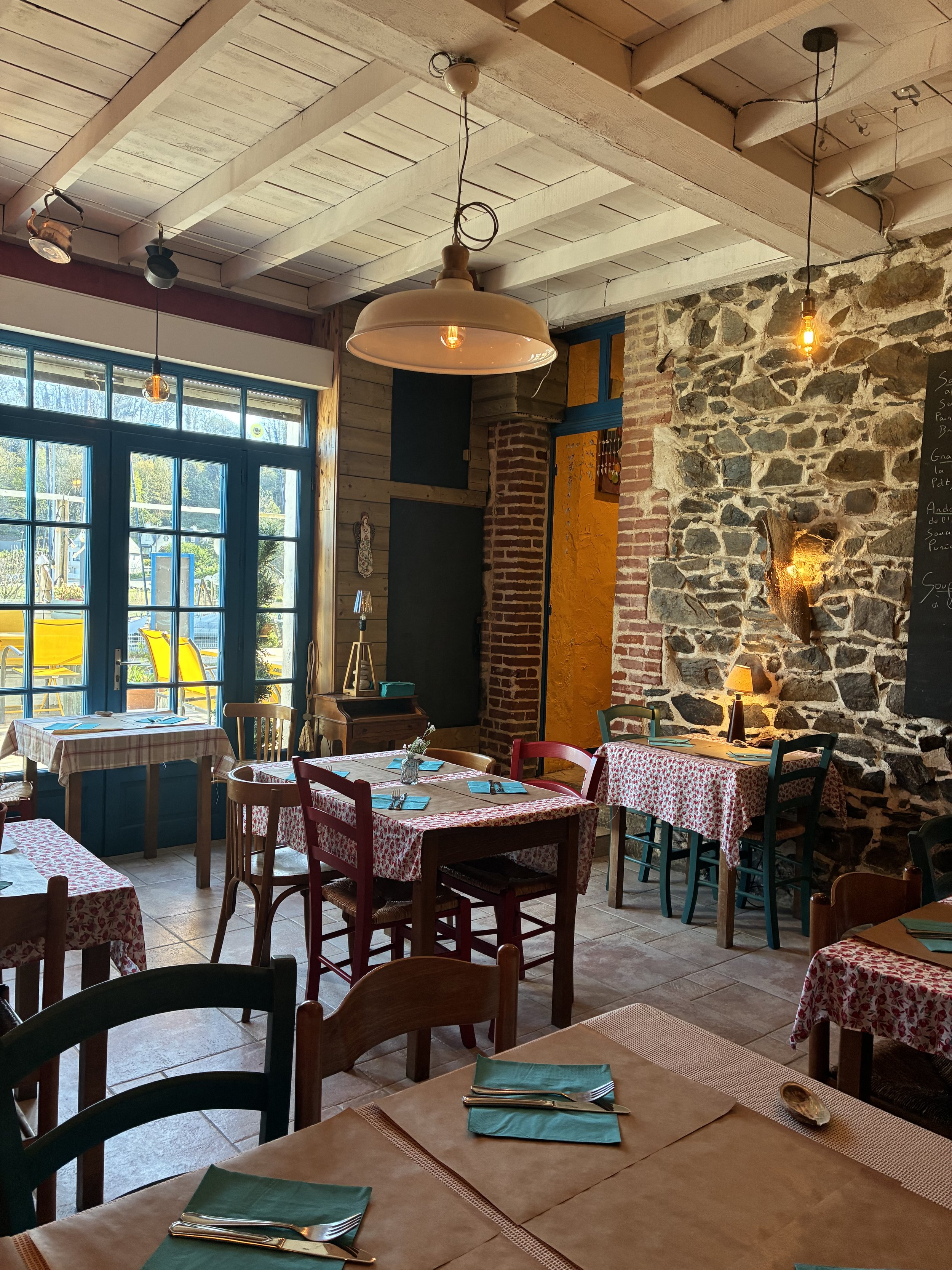 Intérieur d'un restaurant avec des tables décorées de nappes à motifs rouges et blancs, des couverts et des serviettes vertes, murs en pierre et en bois, lampes suspendues avec ampoules visibles, grande porte vitrée avec vue à l'extérieur, ambiance c