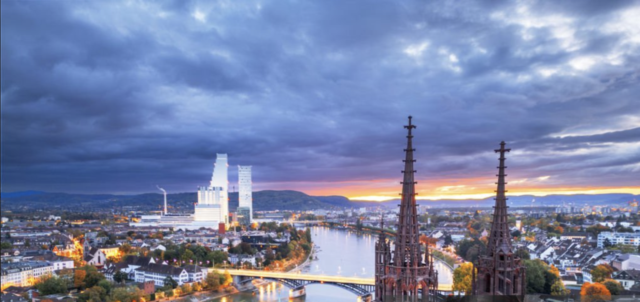Stadtansicht bei Sonnenuntergang mit Wolken, Fluss und Kirchen spitzen im Vordergrund sowie modernen Hochhäusern im Hintergrund.