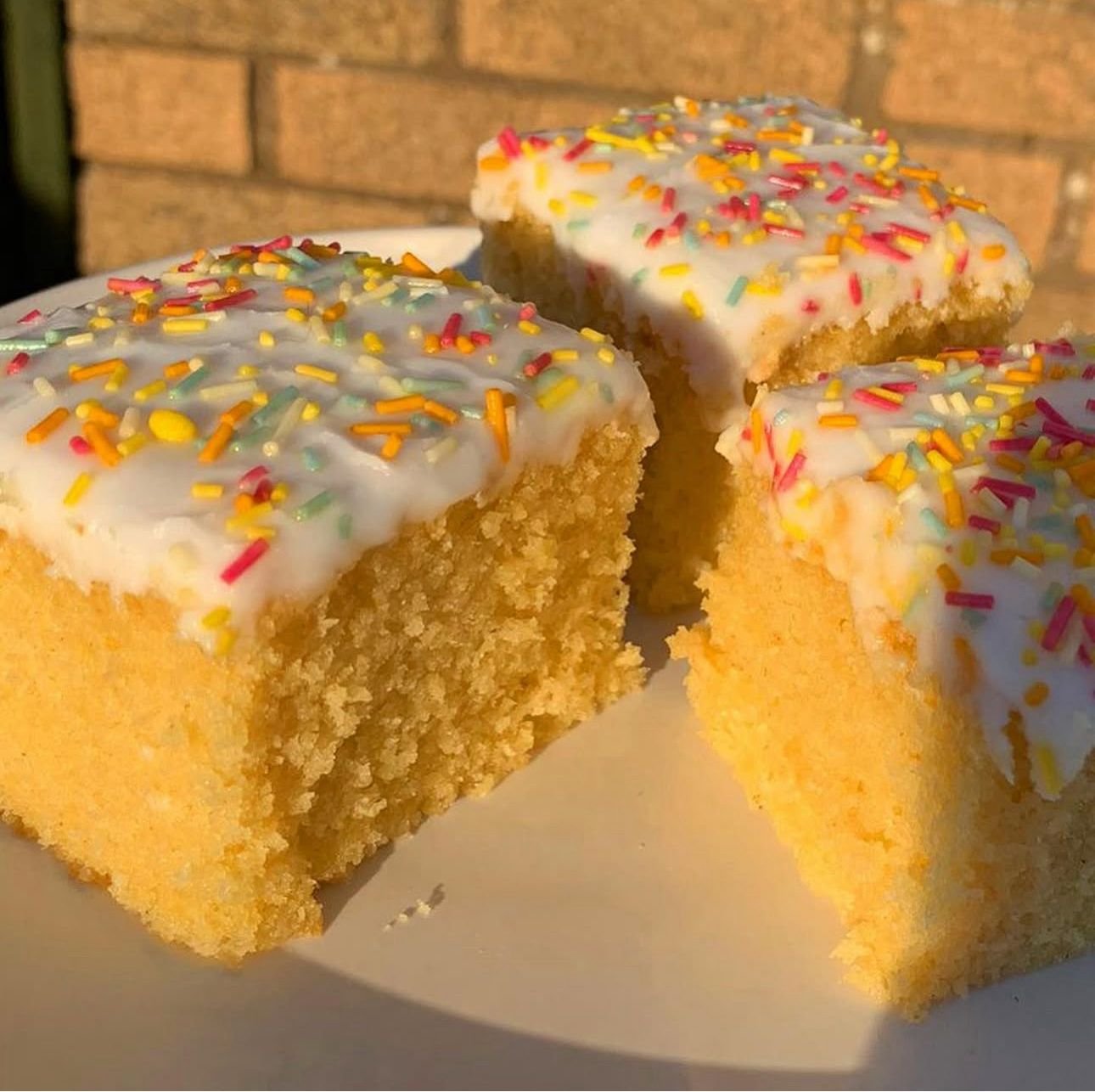 Close-up of two slices of yellow cake with white frosting and colorful sprinkles on top, placed on a white plate with a brick wall background.