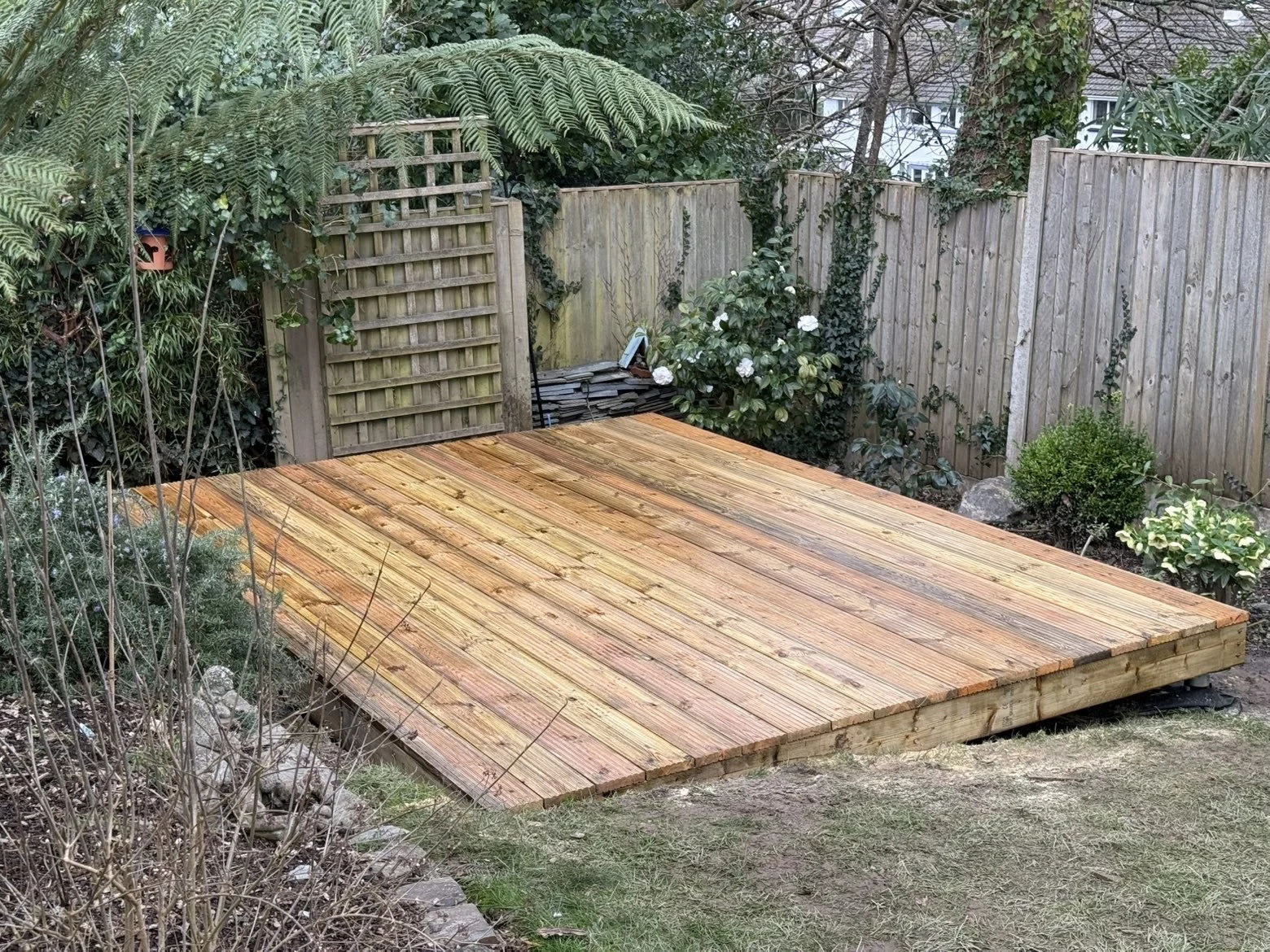 A newly built wooden deck in a backyard garden, surrounded by green plants, bushes, and a wooden fence with a small gate. Some gardening tools and materials are seen in the background.