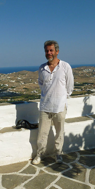 Un homme souriant en chemise blanche et pantalon gris clair, debout sur une terrasse avec vue sur la mer et un paysage rural en arrière-plan.