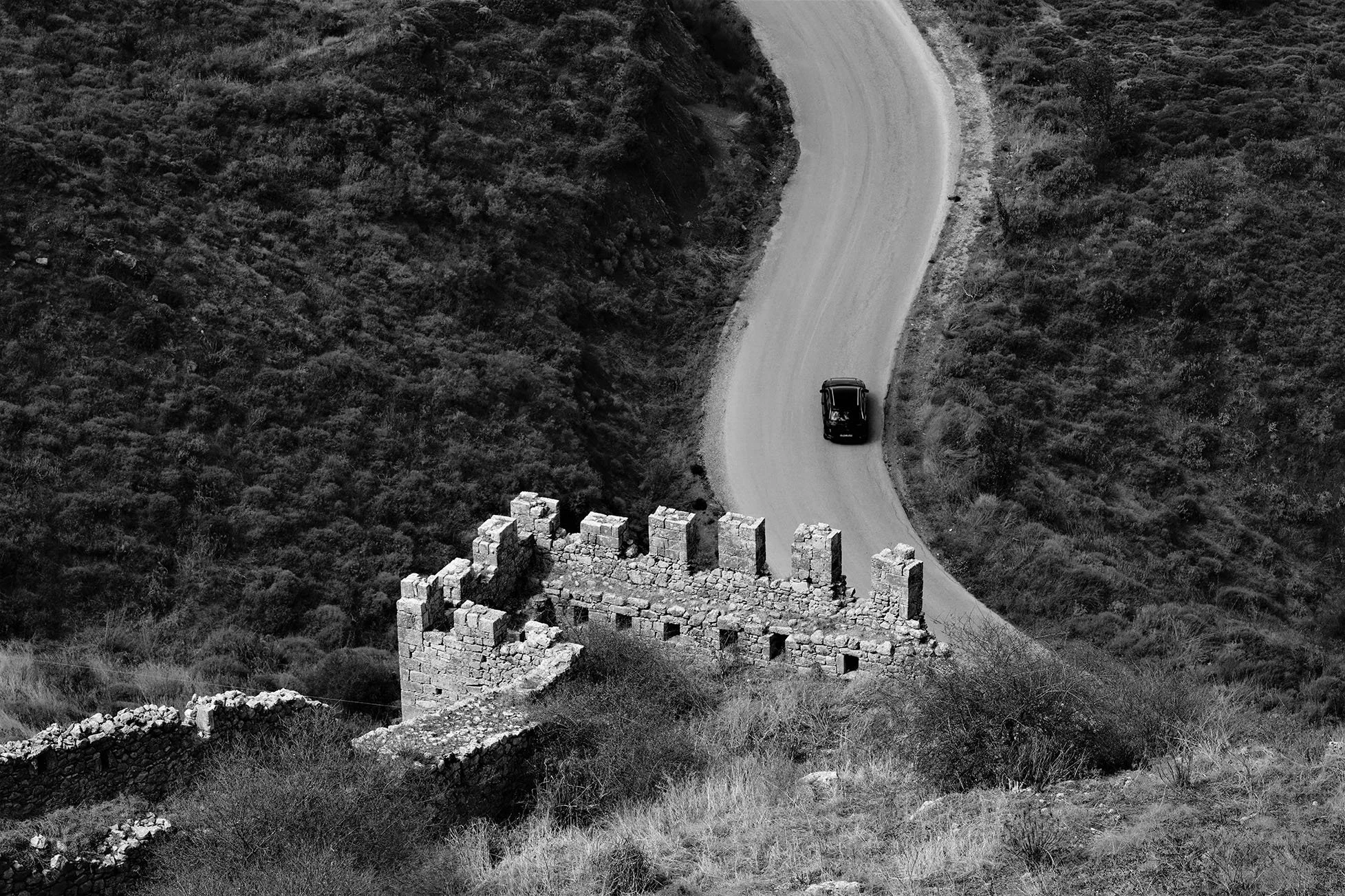 Une route sinueuse traverse un paysage de collines avec une vieille structure en pierre au premier plan. Une voiture noire circule sur la route. L'image est en noir et blanc.