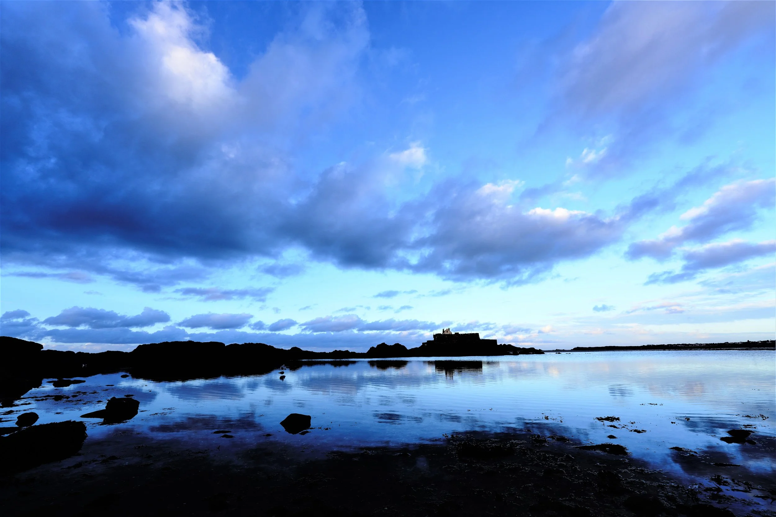 Paysage maritime avec des rochers, un bâtiment au loin et un ciel nuageux reflétant l'eau calme