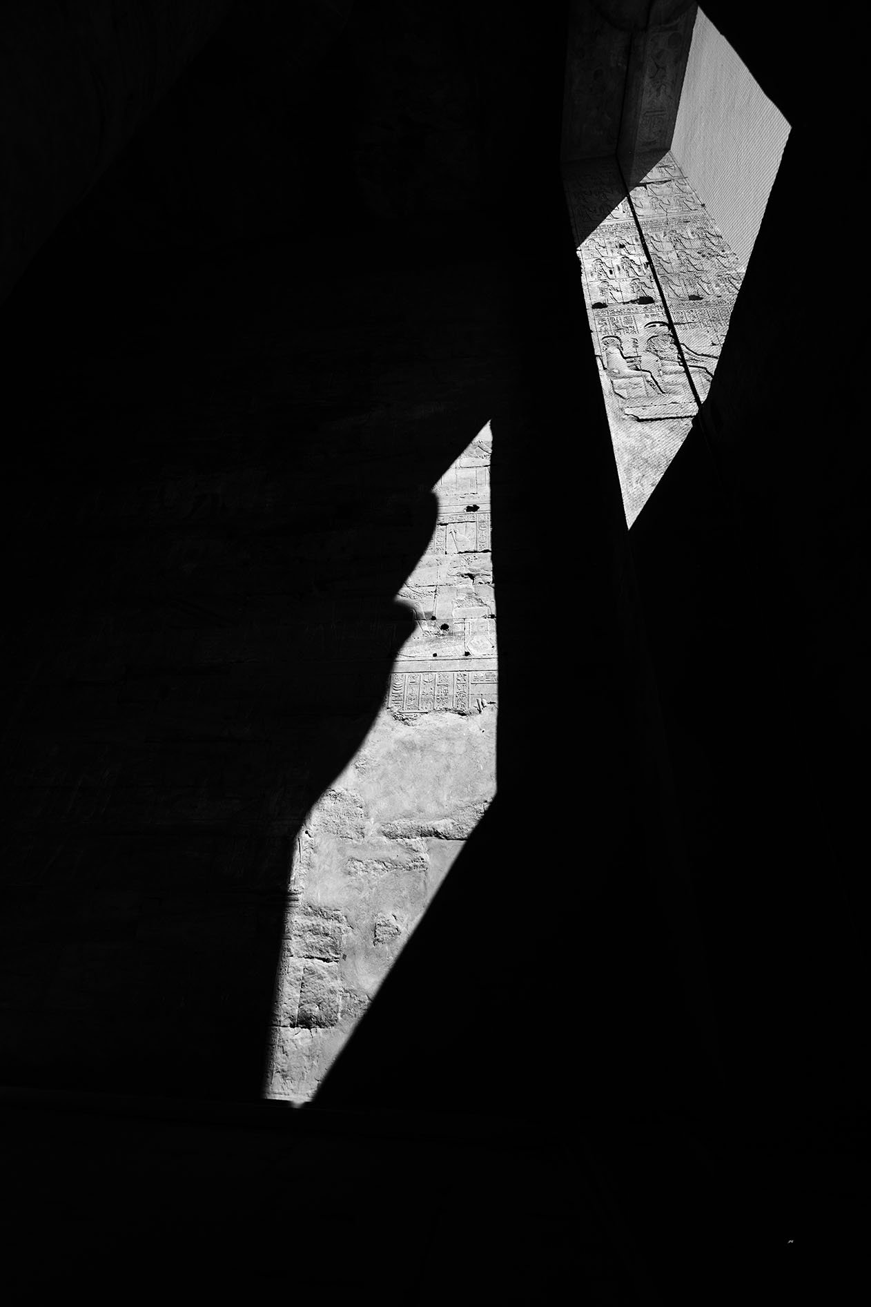 L'image montre une vue en noir et blanc à travers un espace sombre, avec des zones éclairées par la lumière du soleil provenant de deux fenêtres ou ouvertures en haut à droite.