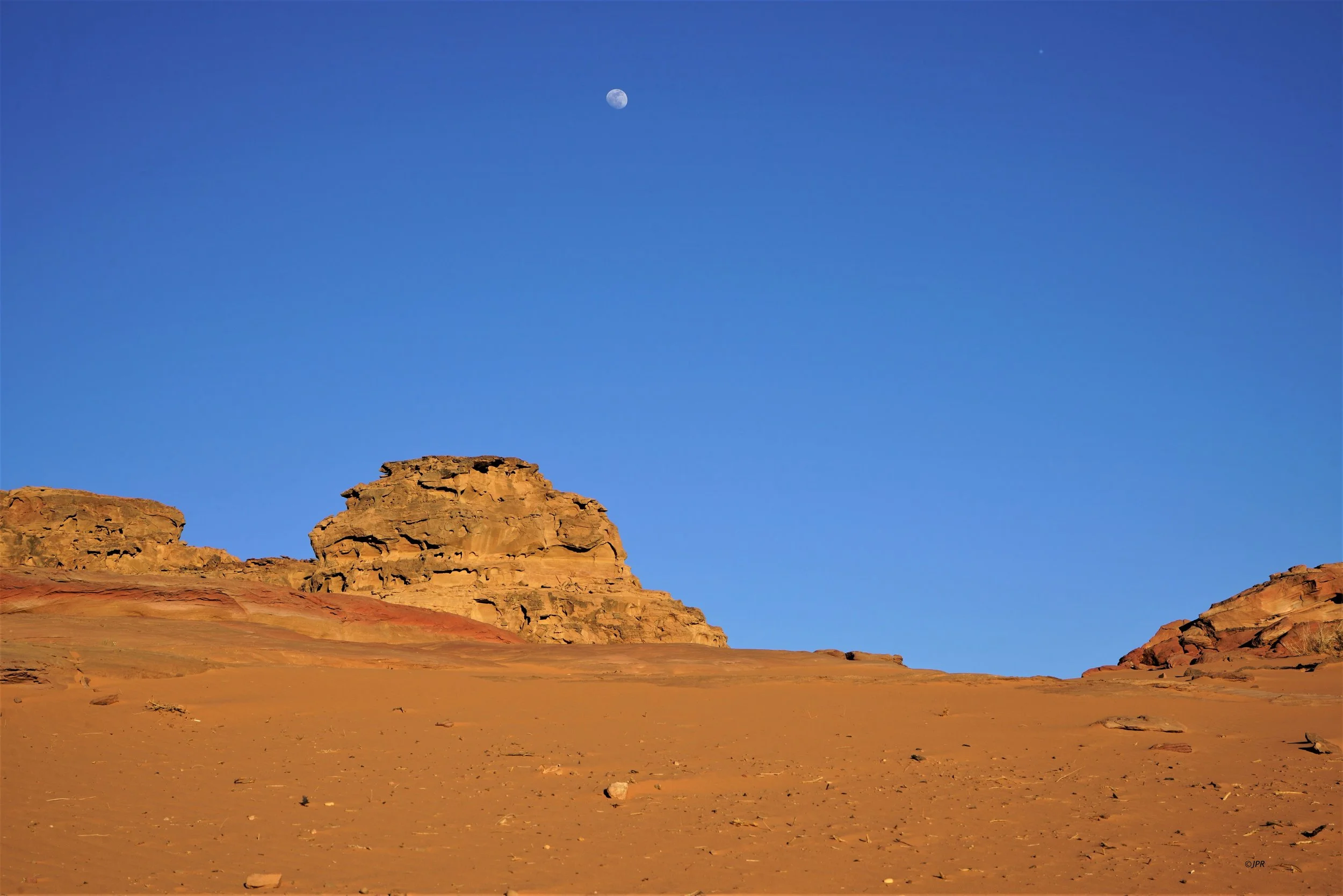 Paysage désertique avec rochers en arrière-plan, ciel bleu clair et lune visible.
