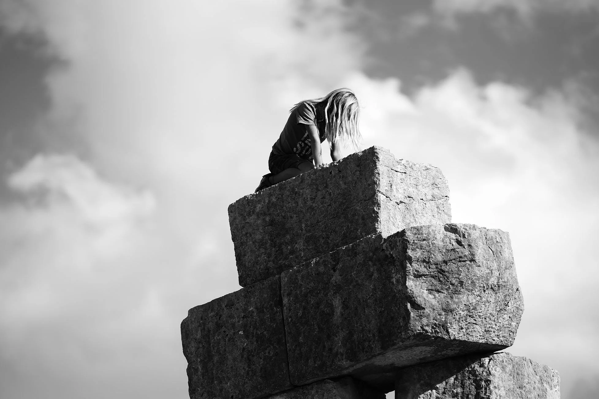 Une jeune fille en t-shirt et pantalon, avec des cheveux longs, regarde vers le bas depuis le sommet d'une pierre ou d'un rocher en équilibre, contre un ciel nuageux.