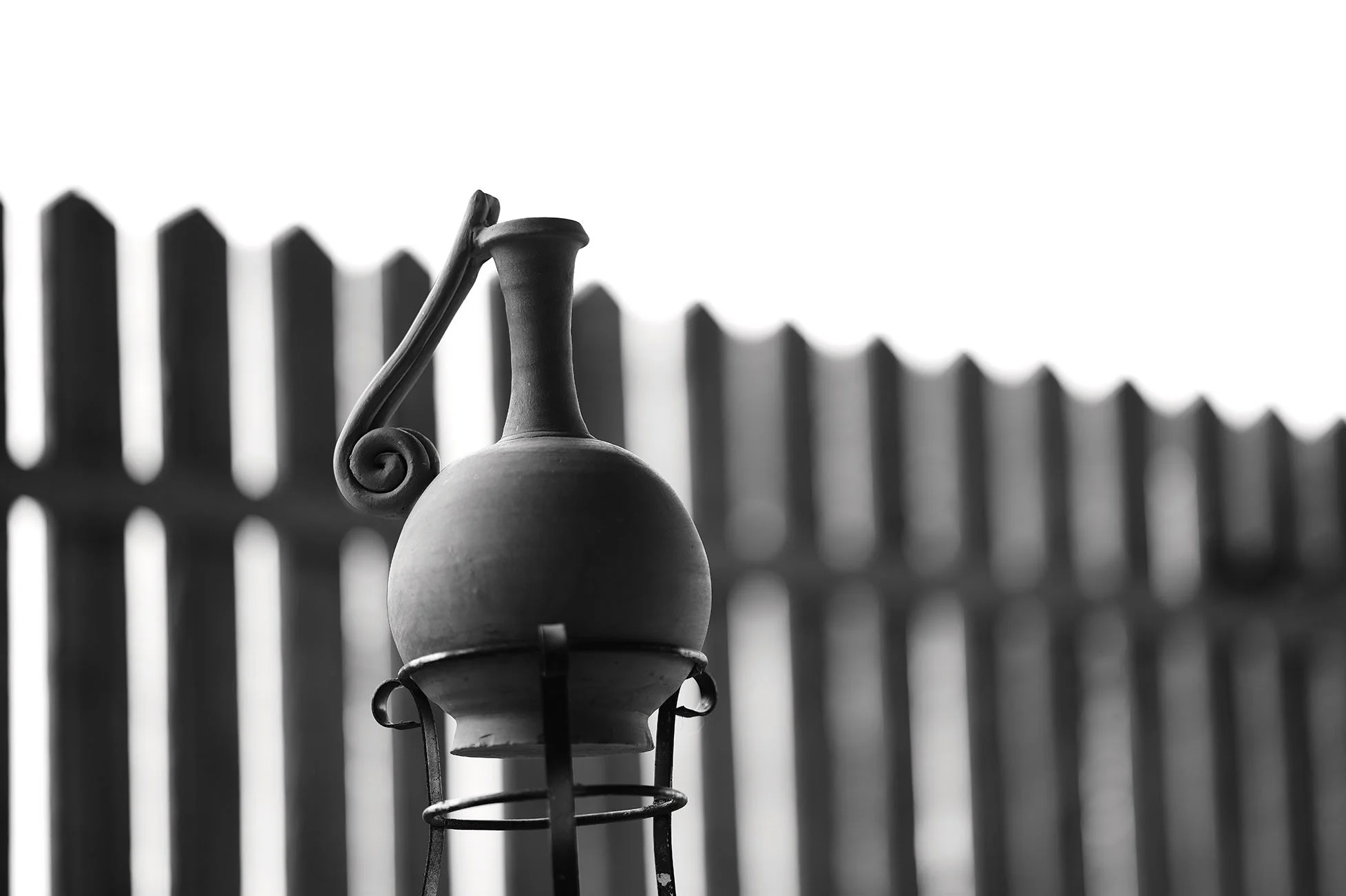 Une lanterne en métal noire, avec un support, devant une clôture en bois en arrière-plan. La photo est en noir et blanc.