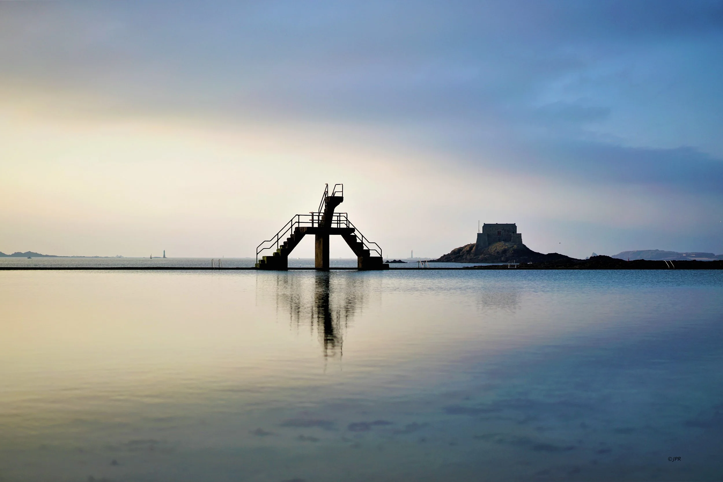 Piscine extérieure avec une plateforme de saut en métal, reflet dans l'eau calme, île avec une forteresse en pierre, ciel nuageux.