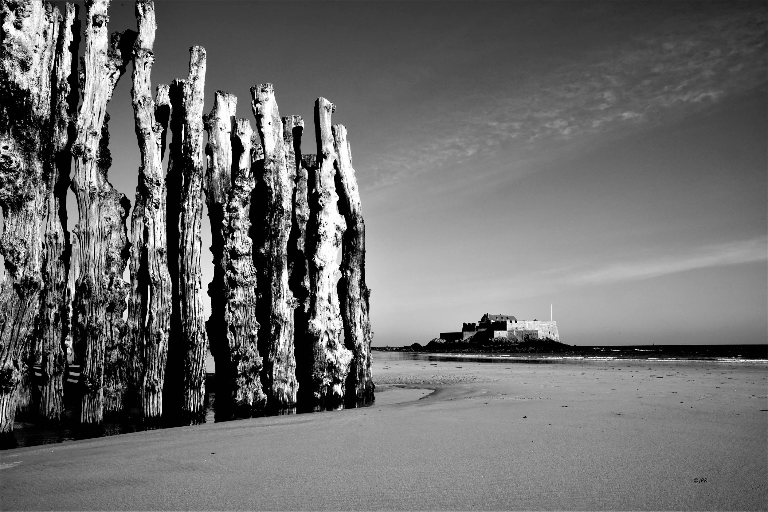 Une plage avec un mur de bois sur la gauche et un fort en arrière-plan, le tout en noir et blanc.