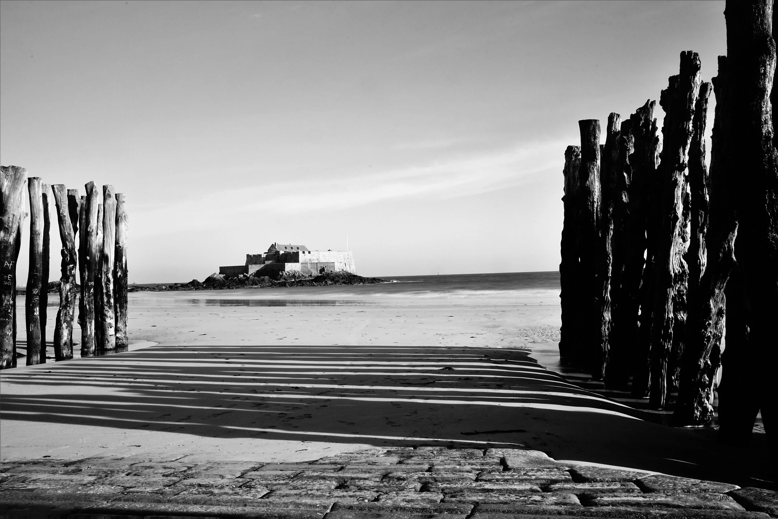Plage avec un mur en pierre au premier plan, des piliers en bois sur les côtés, et un château ou une forteresse à l'horizon, photographié en noir et blanc.