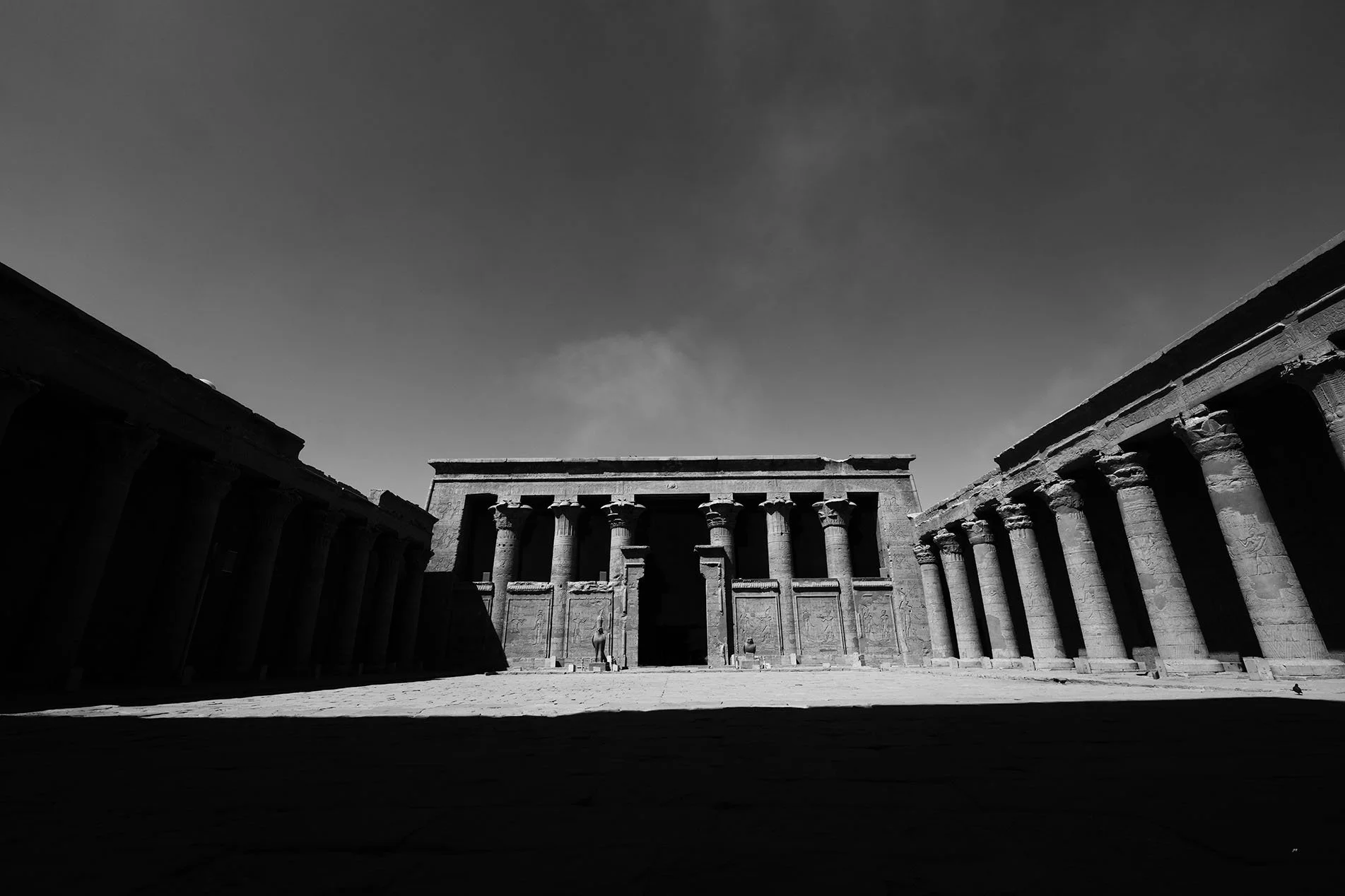 Photographie en noir et blanc d'un ancien site architectural avec des colonnes et une façade centrale, sous un ciel clair.