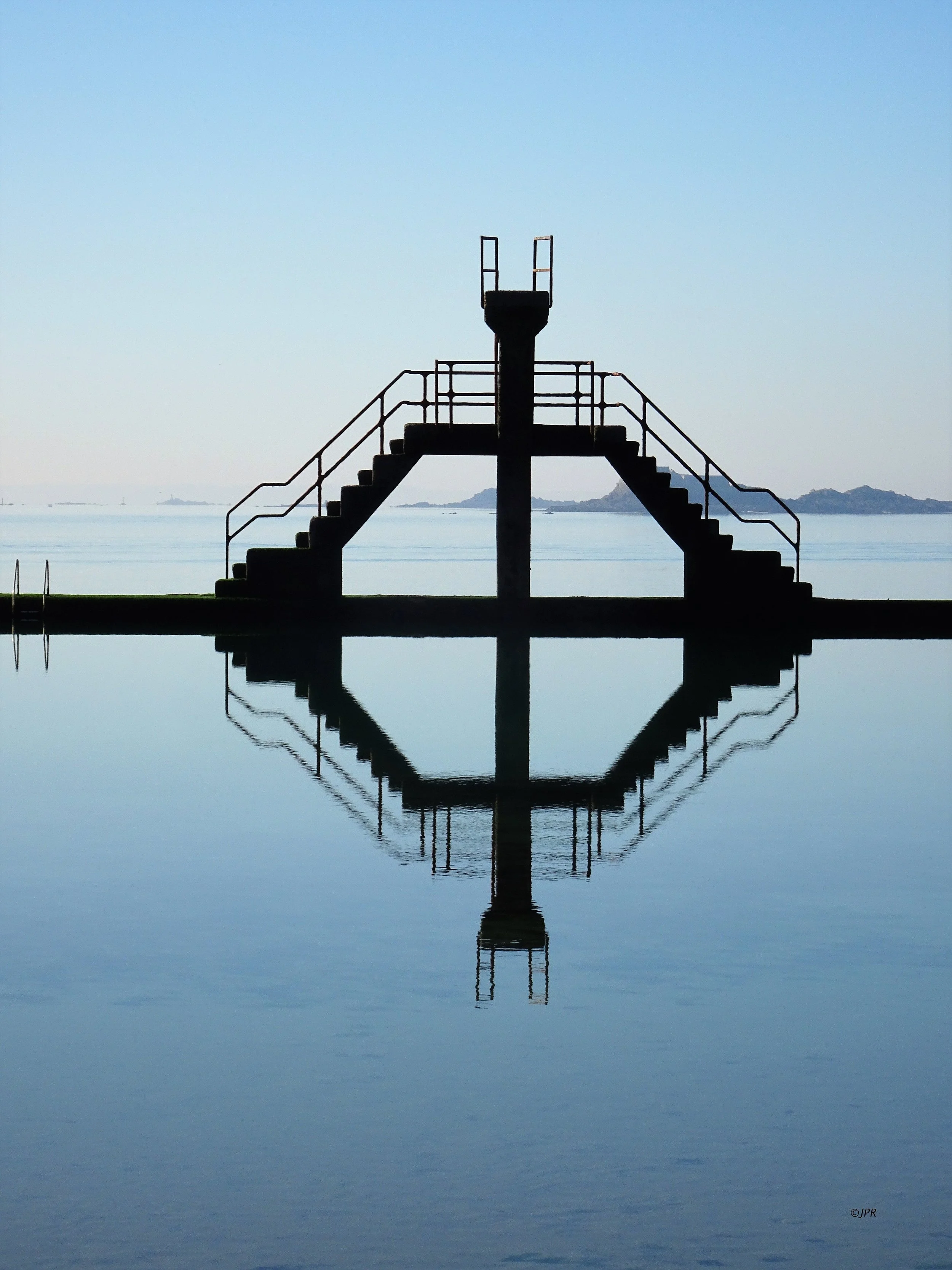Silhouette d'une plateforme de baignade avec deux escaliers en spirale menant à une plateforme centrale, reflet dans l'eau calme, avec un ciel dégagé et la mer en arrière-plan.