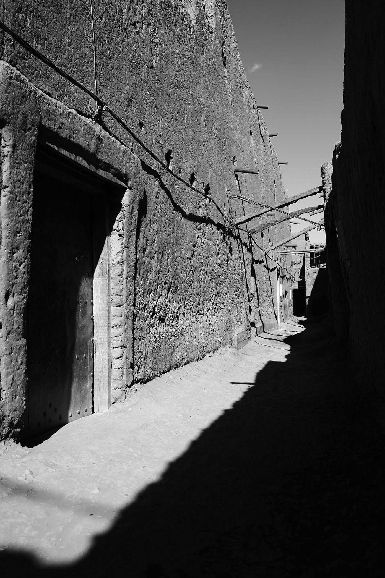 Une ruelle étroite avec des murs en pierre, des câbles électriques suspendus, et un sol en terre, en noir et blanc.