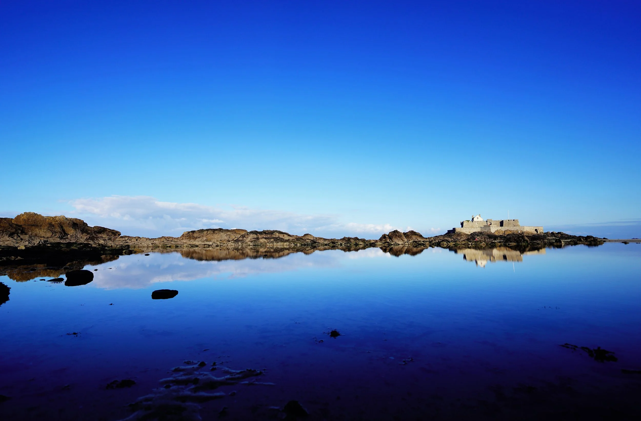 Un paysage côtier avec une mer calme, des rochers, un bâtiment en pierre, et un ciel bleu avec quelques nuages.