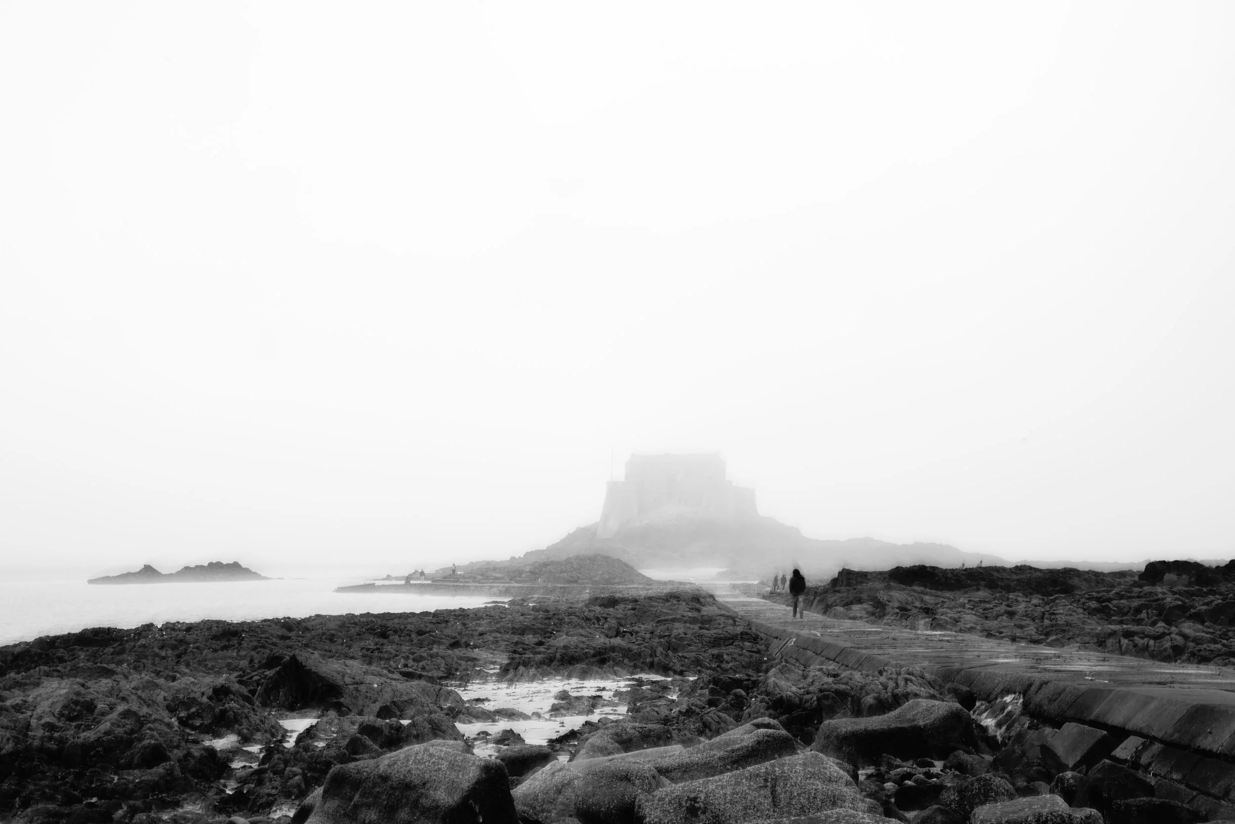 Paysage côtier en noir et blanc avec un château en haut de la colline en arrière-plan, rochers au premier plan et deux personnes marchant vers le château.