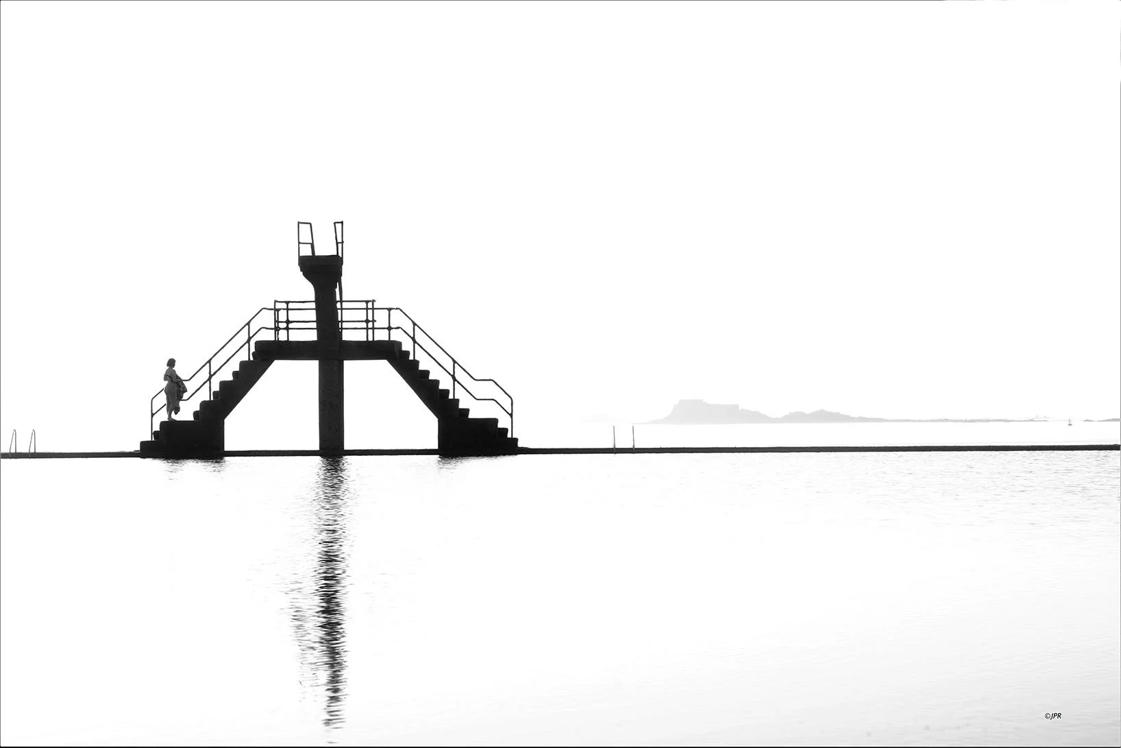 Un plongeoir de piscine en silhouette avec une personne qui marche sur le côté, reflet dans l'eau calme, horizon extérieur minimaliste.