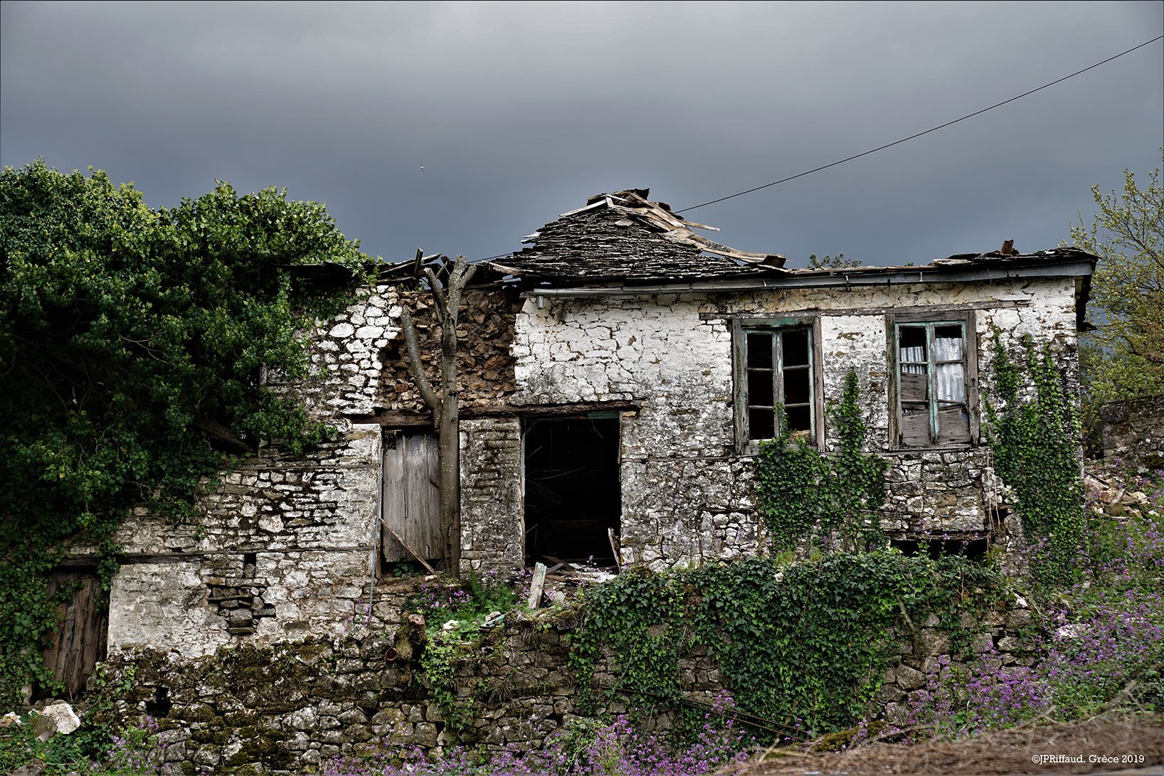 Maison abandonnée en pierre avec fenêtres cassées, entourée de végétation, sous un ciel nuageux.