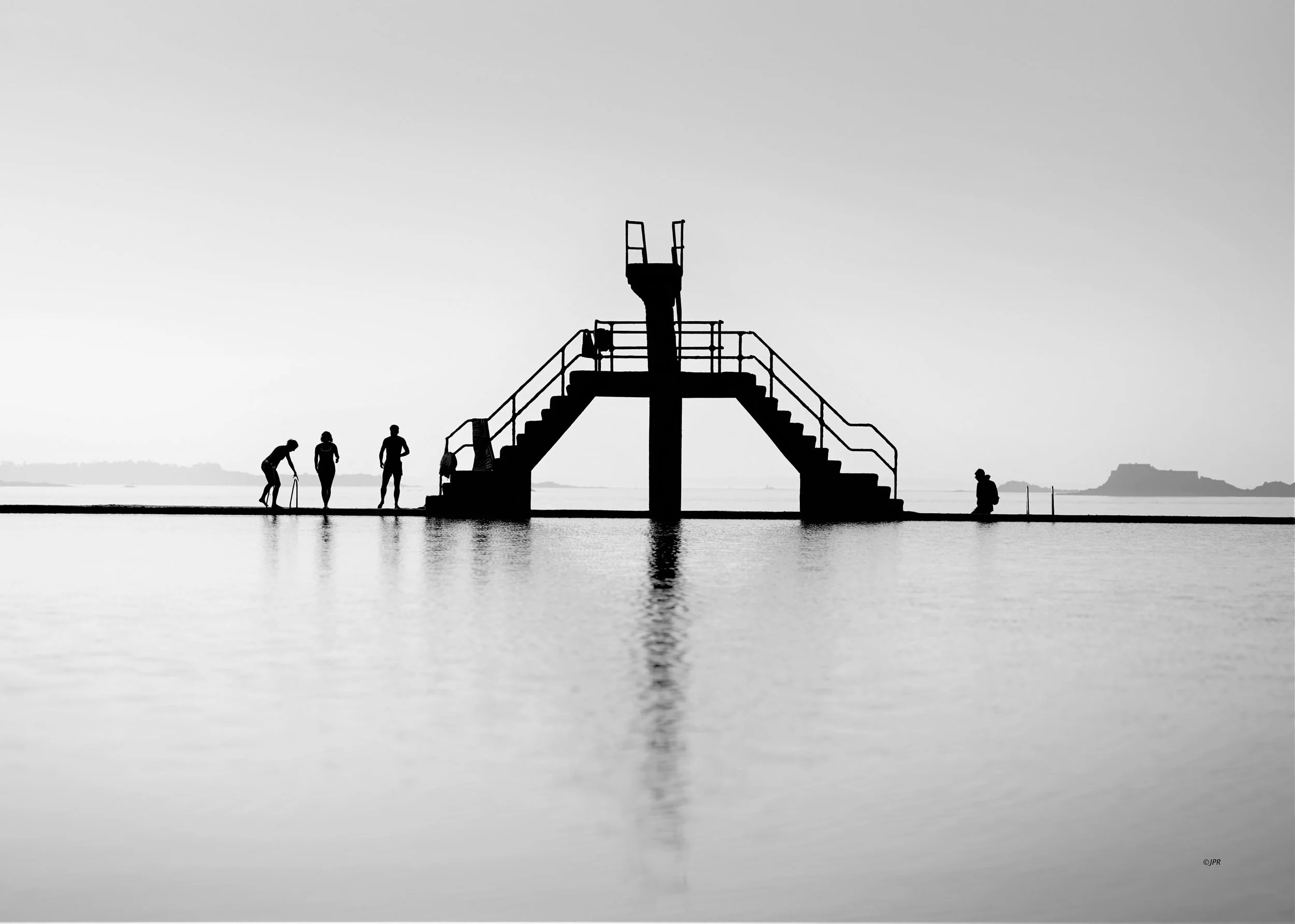 Silhouettes de quatre personnes près d'une structure de plongée surplombant l'eau, avec un horizon et une île en arrière-plan, en noir et blanc.