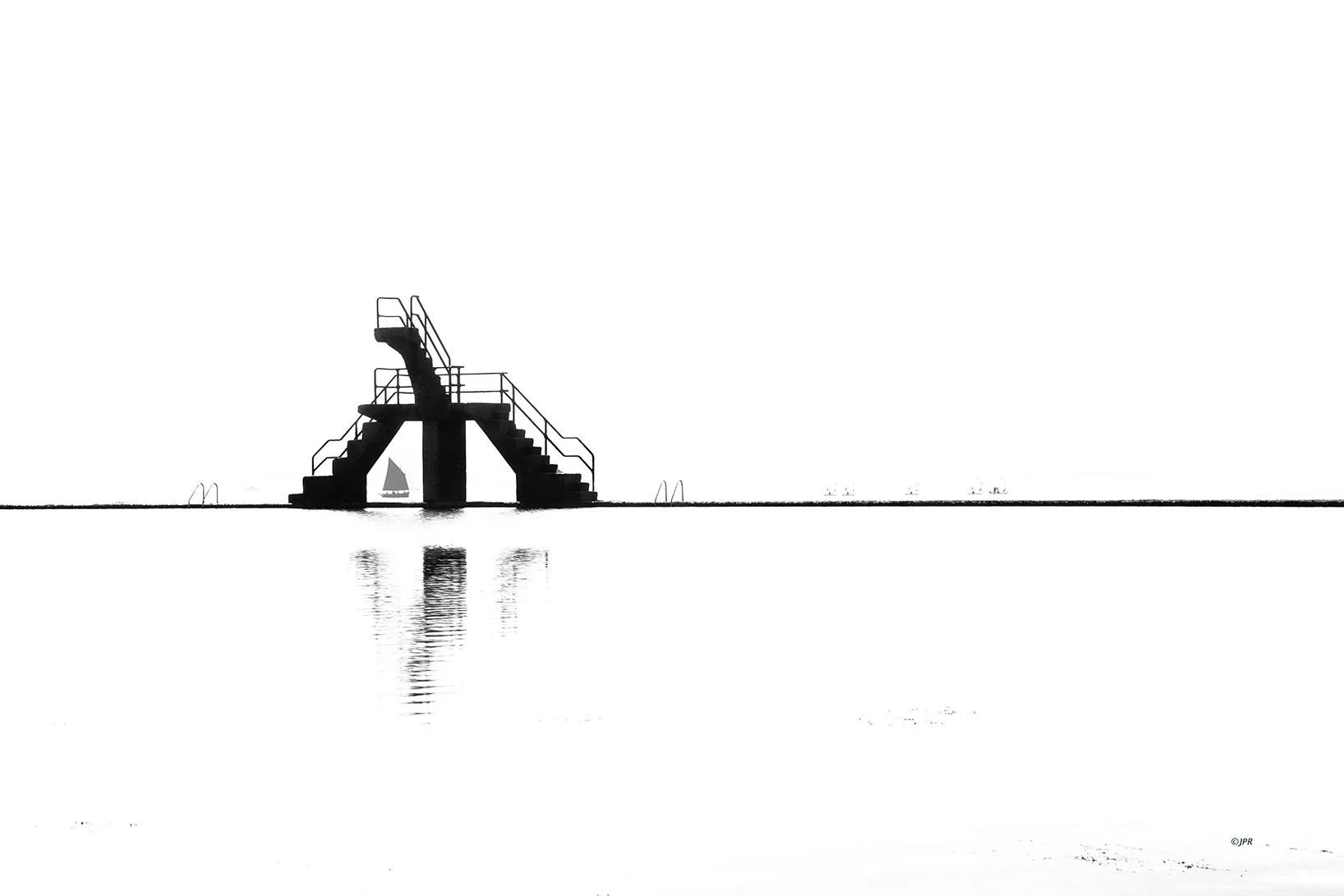Structure de plongée avec un escalier en acier sur une plage, reflet dans l'eau, bateau à voile au loin, fond blanc minimaliste.