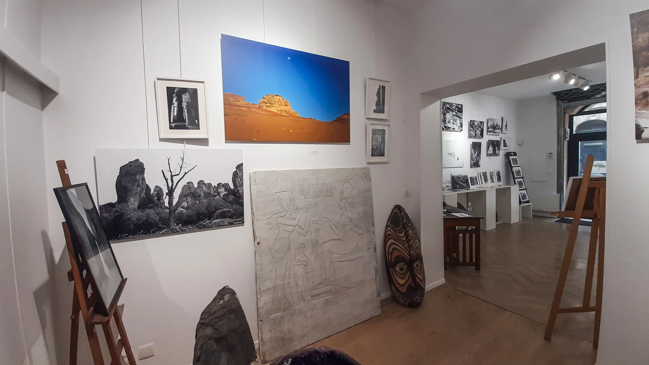 Une galerie d'art avec plusieurs tableaux, photographies et œuvres sculpturales exposés sur des murs blancs, avec un sol en bois et un éclairage ciblé sur les œuvres.