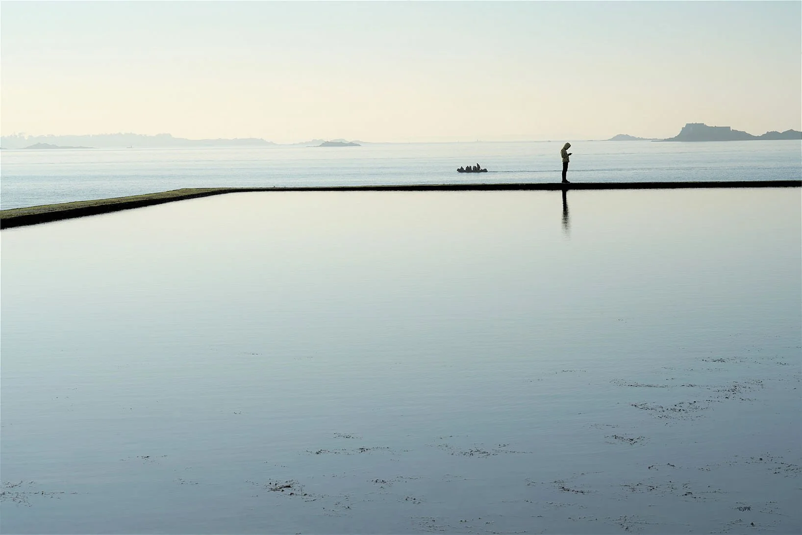 Un homme seul se tient sur une falaise au bord de la mer, regardant son téléphone. La mer calme reflète le ciel clair et il y a des îles lointaines en arrière-plan.