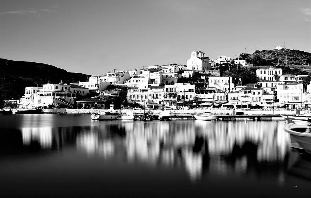 Photographie en noir et blanc d'une ville côtière avec des bâtiments blancs sur une colline et un port avec des bateaux à la surface de l'eau calme.