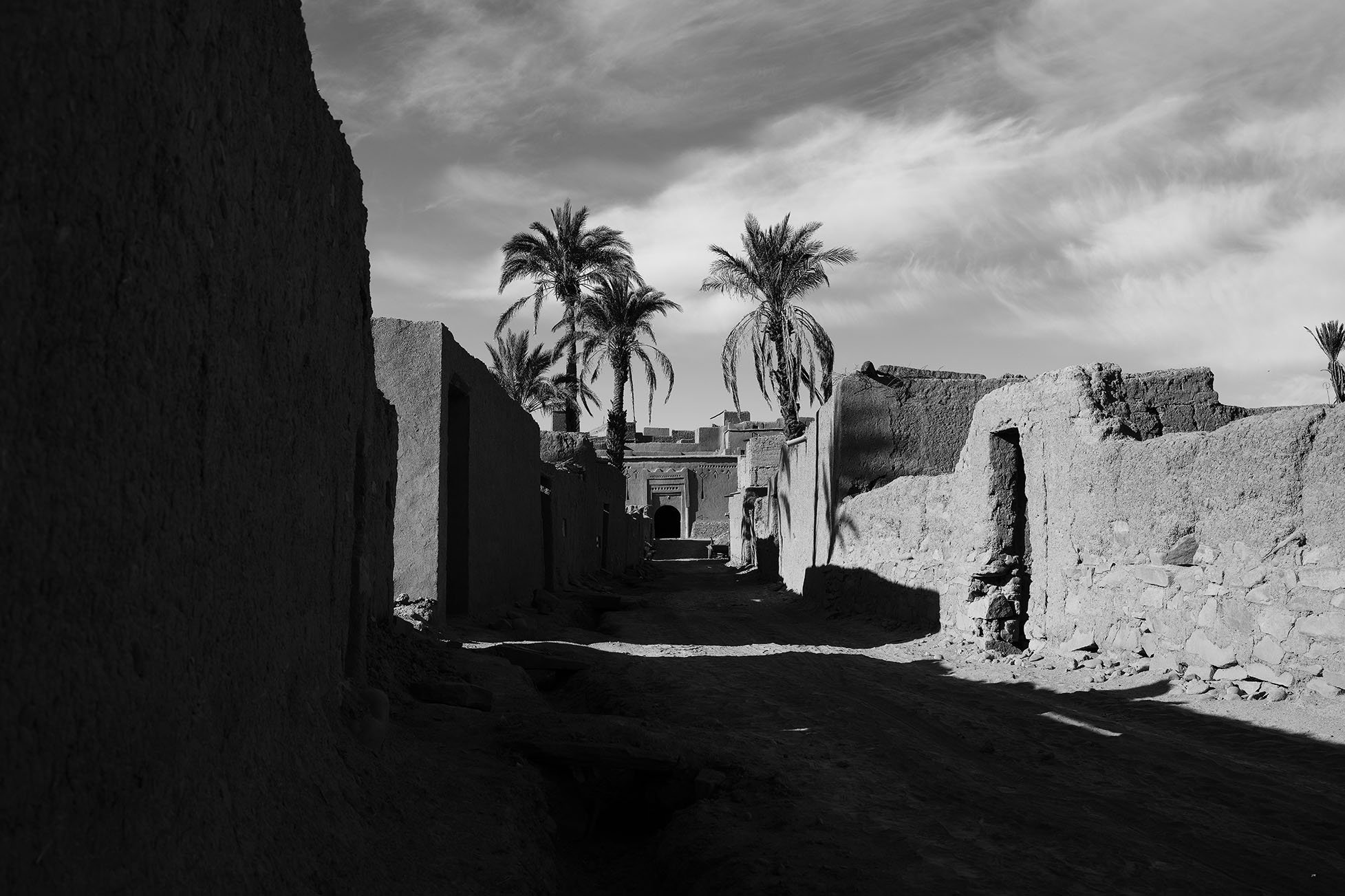 Une rue désertique dans une vieille ville avec des murs en terre cuite, des palmiers au fond et un ciel nuageux.