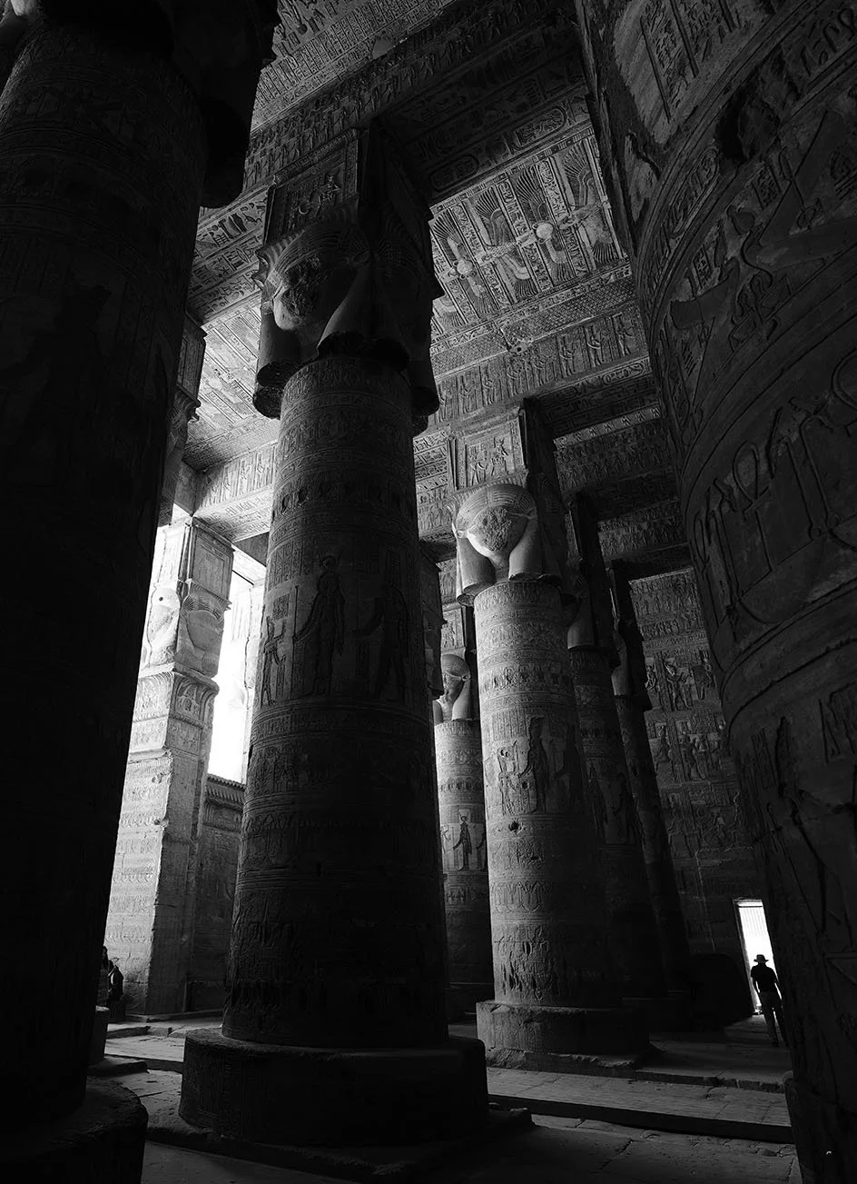 Intérieur d'un temple égyptien ancien avec des colonnes ornées de hiéroglyphes et de statues de figures humaines, en noir et blanc.