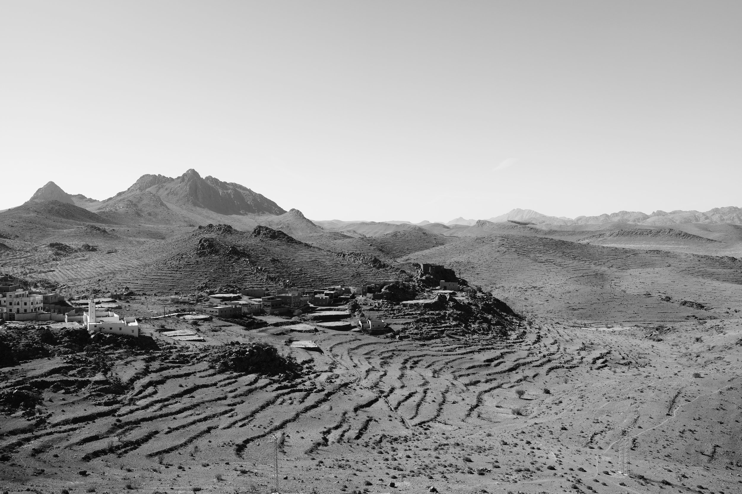 Paysage désertique avec petites maisons blanches, montagnes en arrière-plan et terrasses agricoles en premier plan, en noir et blanc.
