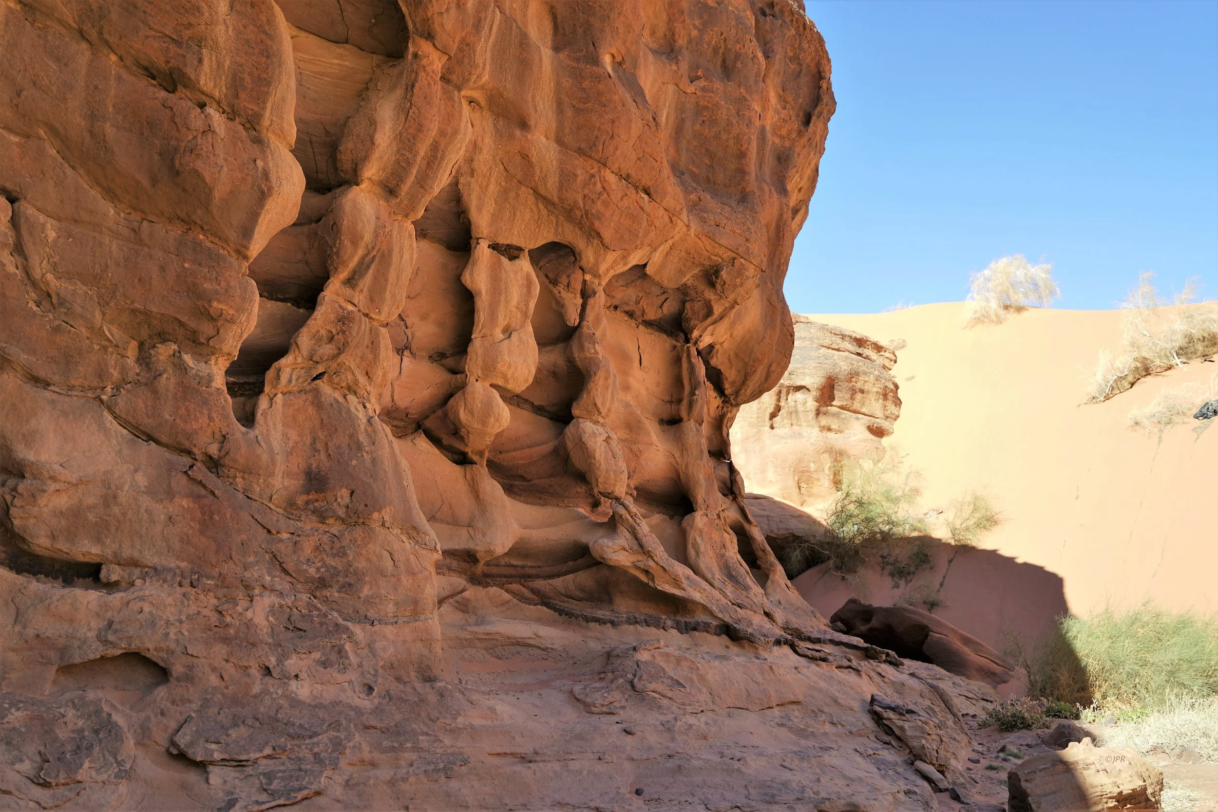Rochers de couleur rouge dans un désert avec une dune en arrière-plan et un ciel bleu clair.