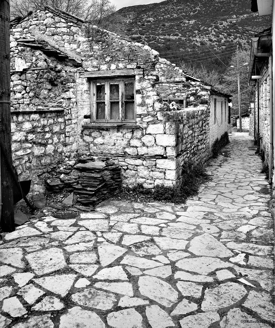 Une vieille maison en pierre sur une rue pavée dans un village de montagne, avec un paysage de collines en arrière-plan