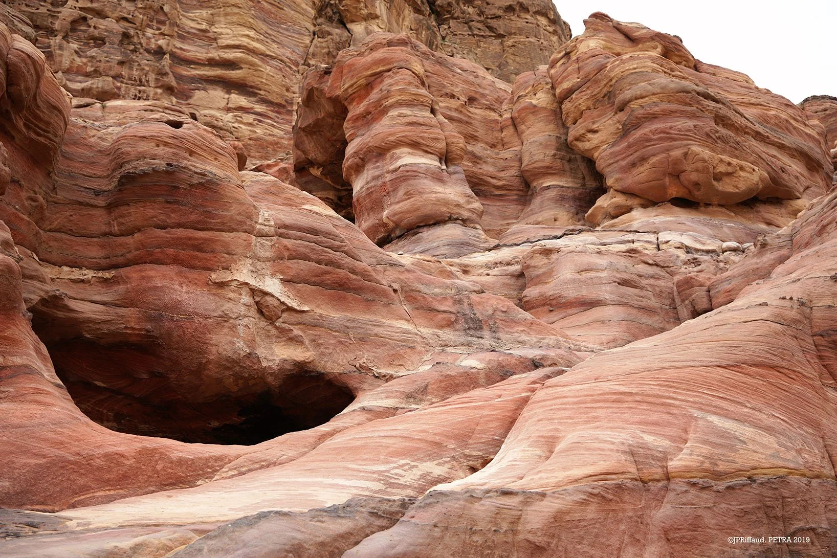 Formation rocheuse colorée rouge, rose et beige avec des couches stratifiées, dans un canyon aride, ciel partiellement visible en haut.