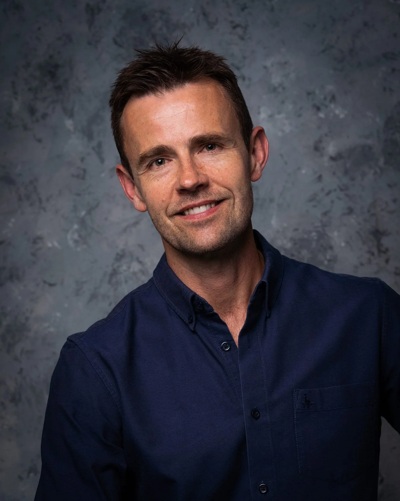Portrait of a young man with dark hair, wearing a navy blue button-up shirt, smiling against a textured gray background.