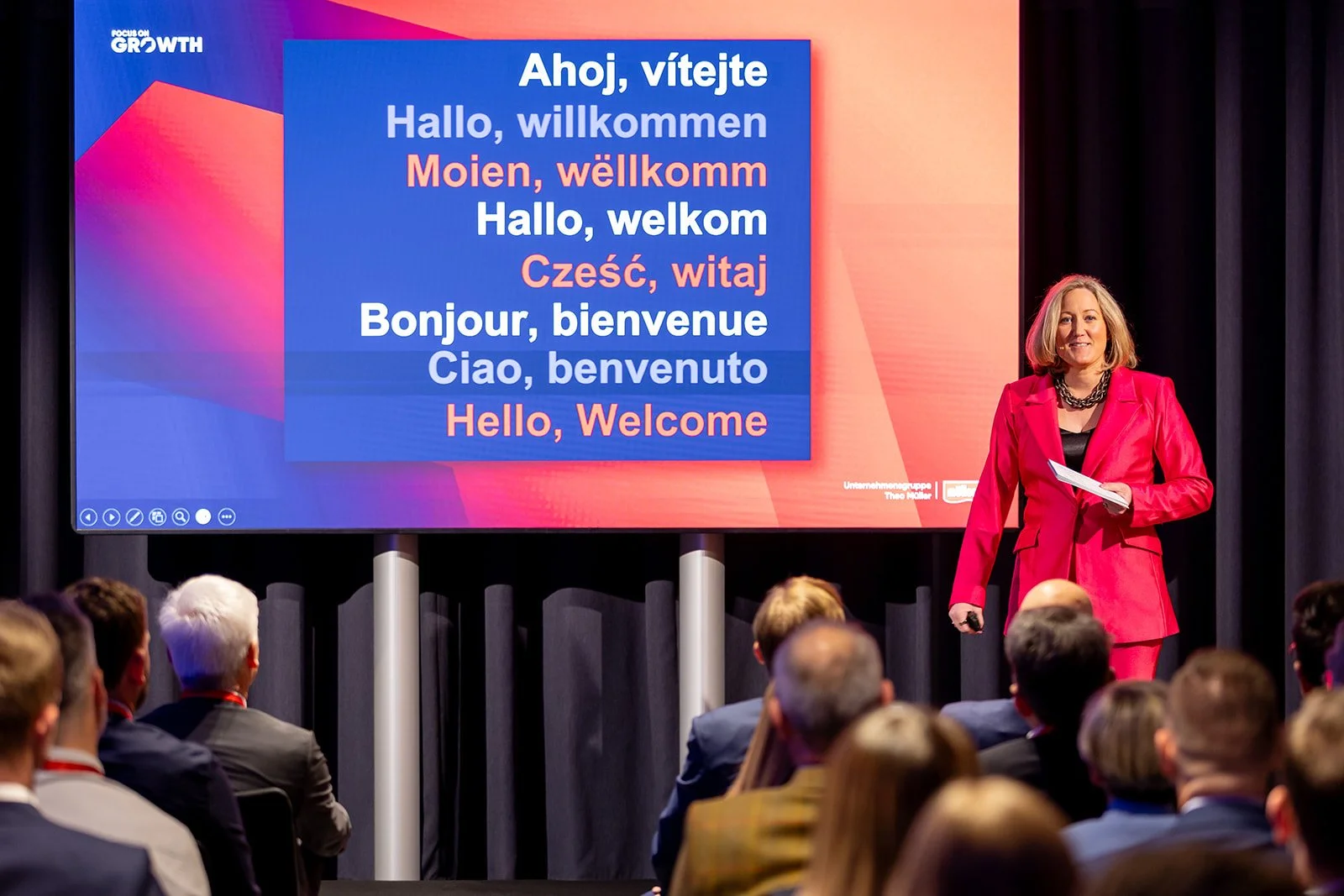 A woman in a pink suit giving a presentation on stage, with a large screen behind her displaying 'Hello, Welcome' greetings in multiple languages.