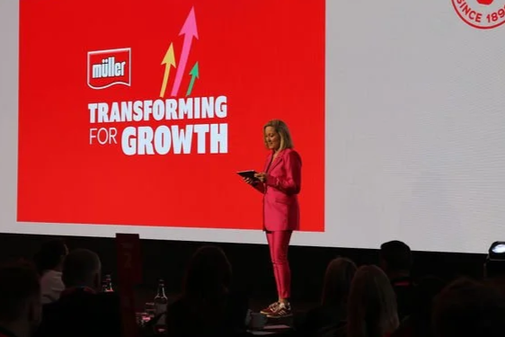 A woman in a pink suit presenting on stage in front of a large screen displaying the Müller logo and the slogan 'Transforming for Growth' with upward arrows.