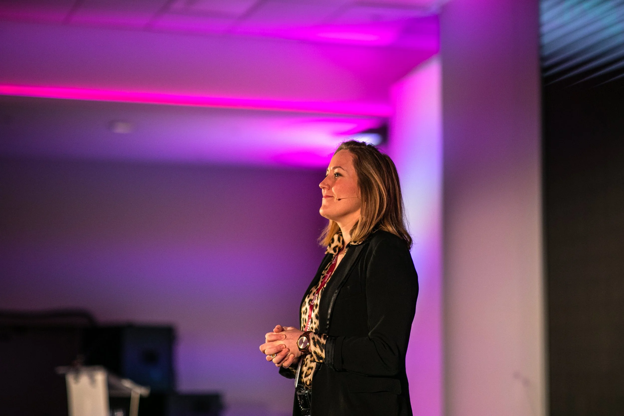 A woman with shoulder-length hair wearing a black blazer and leopard print blouse, standing with her hands clasped, speaking at an event with purple and pink lighting in the background.