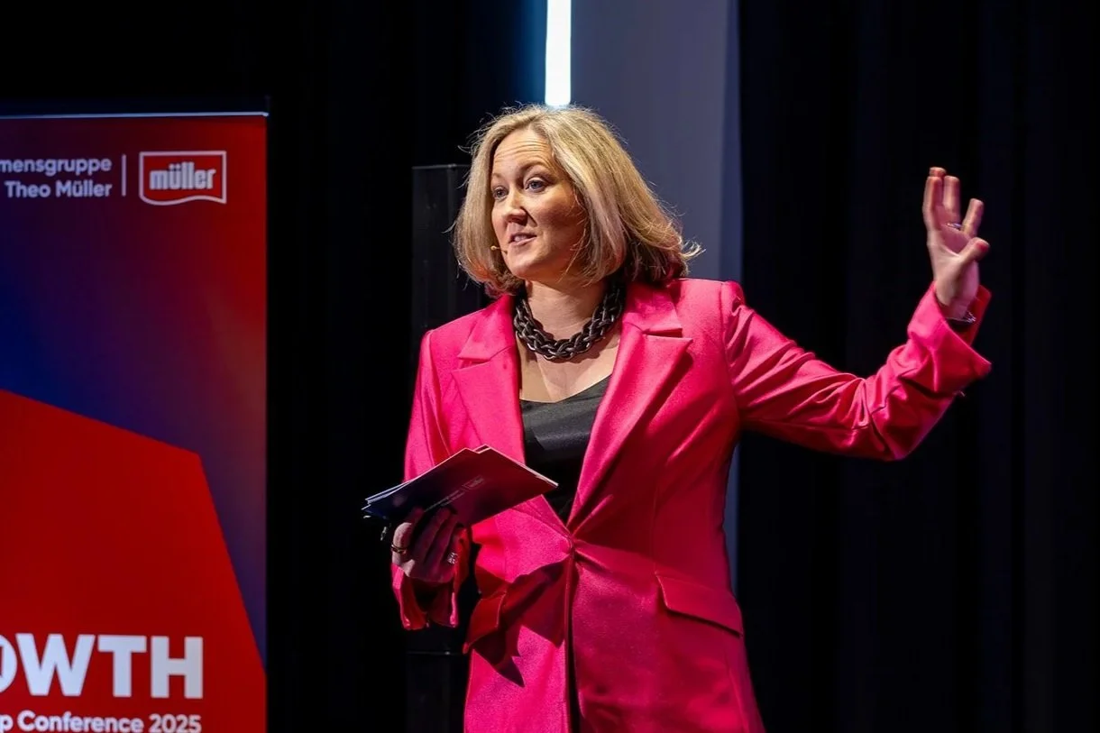 A woman in a bright pink blazer speaking at a conference, holding some notes, with a backdrop displaying the logos for Mäntsä and the event.