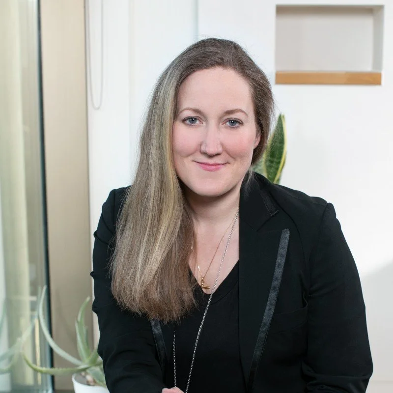 A woman with long blonde hair smiling, wearing a black blazer and necklaces, sitting indoors with a plant and shelves in the background.